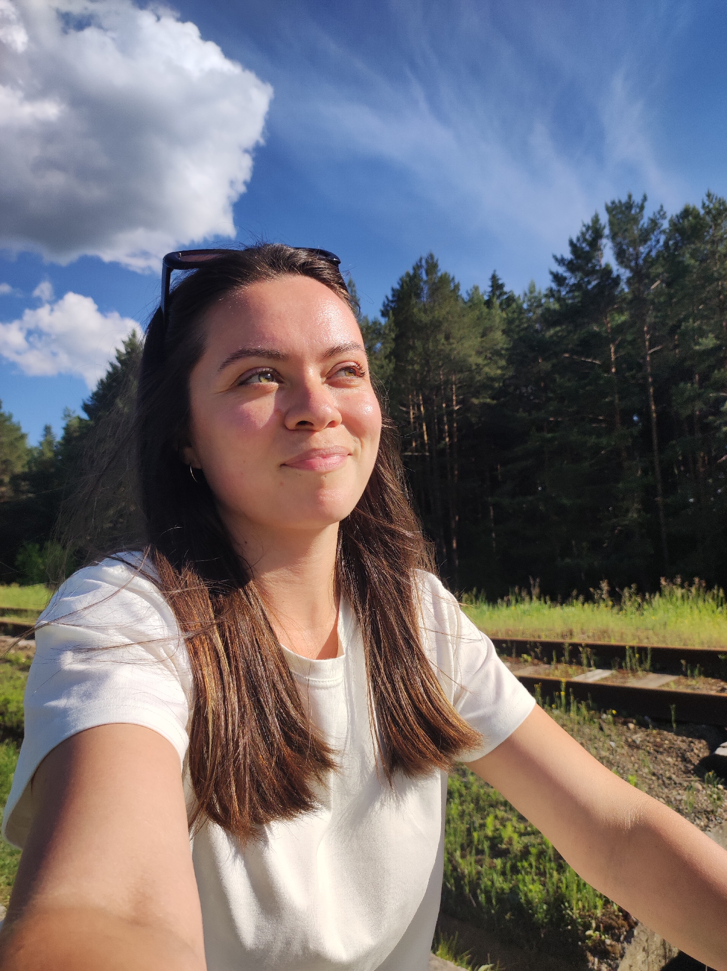 A young woman with long brown hair and a white t-shirt is taking a selfie outdoors. Sunglasses are perched on top of her head. She is looking up and to her right, with a slight smile. The background includes a blue sky with white clouds, a dense green forest, and a set of train tracks with surrounding vegetation. The location may be in or near the city of Chaikovsky, Russia, based on implied location context.