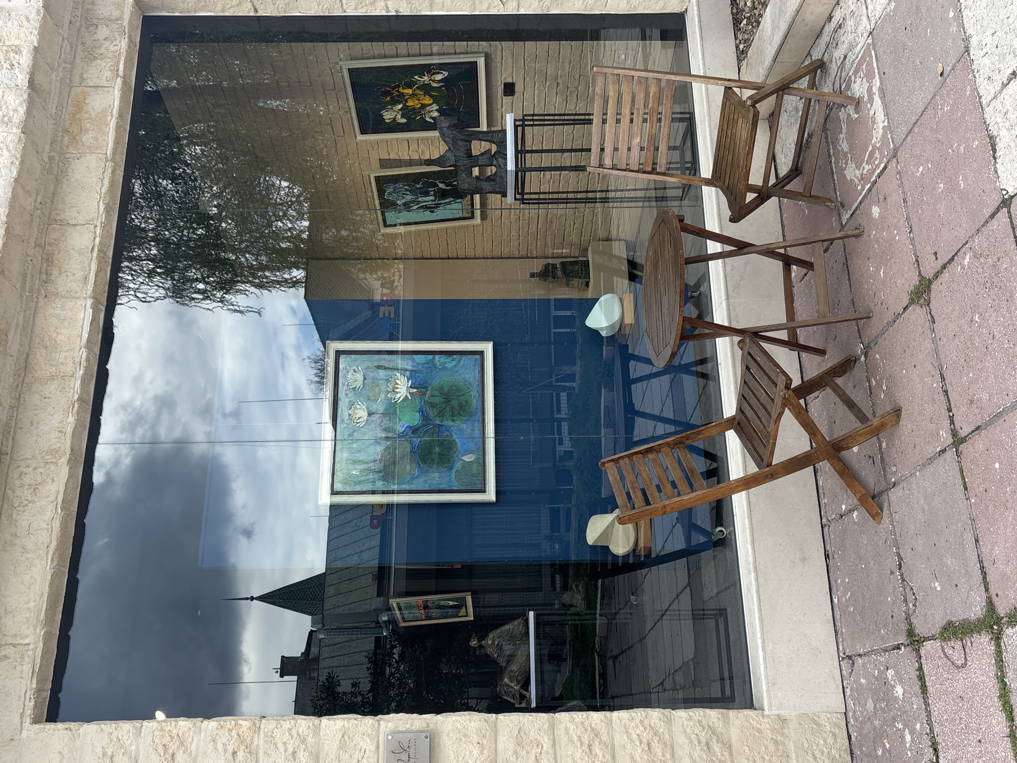The image depicts a storefront window in Varna, Bulgaria. The reflection in the window shows a cloudy sky, a building with a spire, and trees. The interior of the store, visible through the window, displays paintings, sculptures, and furniture. Two wooden folding chairs and a small round wooden table sit on the sidewalk outside the store. The sidewalk is paved with pinkish-red tiles.