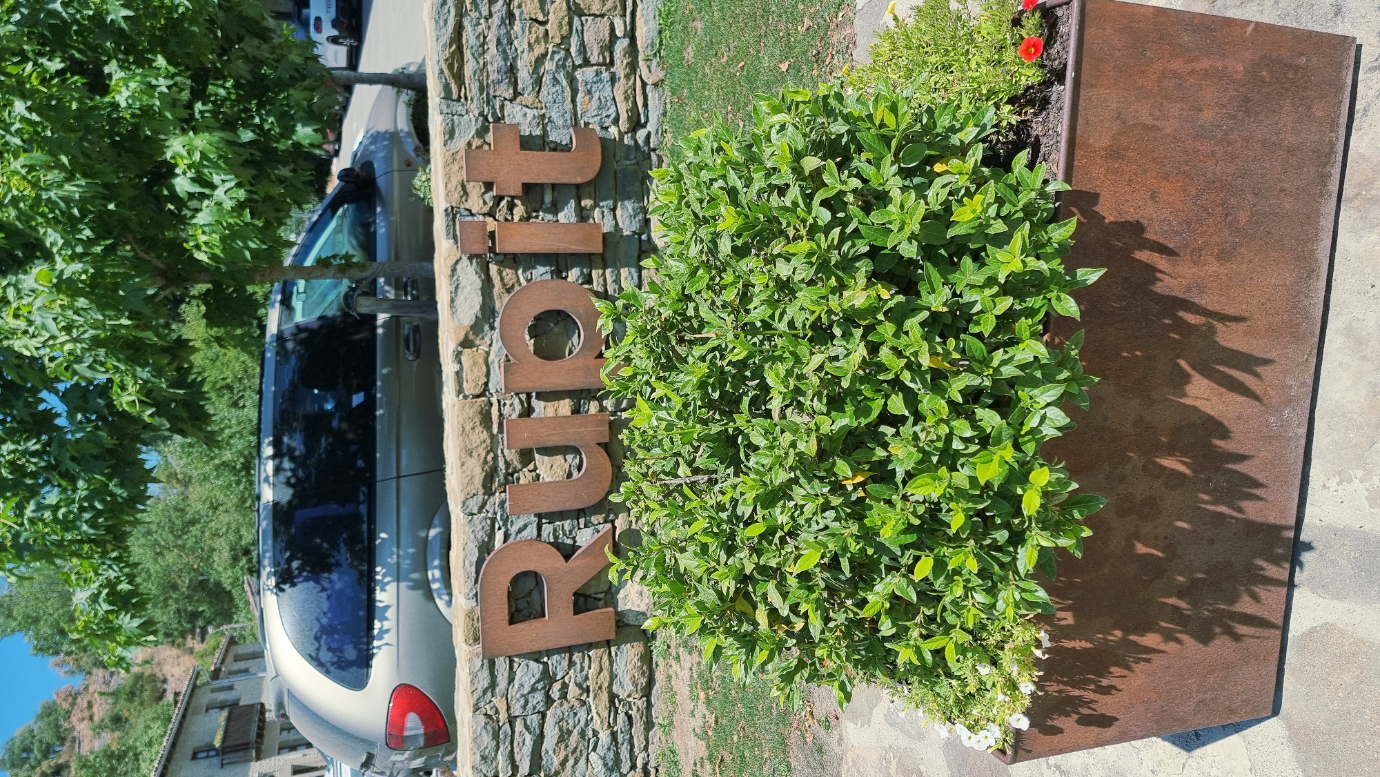 The image shows a sign with the word "Rupit" made of rusty metal letters attached to a stone wall. In front of the sign is a large planter box filled with lush green bushes and some small white flowers. To the left and behind the bushes, a tan minivan is parked. A building with a balcony and a tiled roof is visible in the background, nestled into a hillside. Trees with green leaves are also present, casting shadows on the ground. The ground appears to be paved with stone slabs. The scene is outdoors, under a clear blue sky.