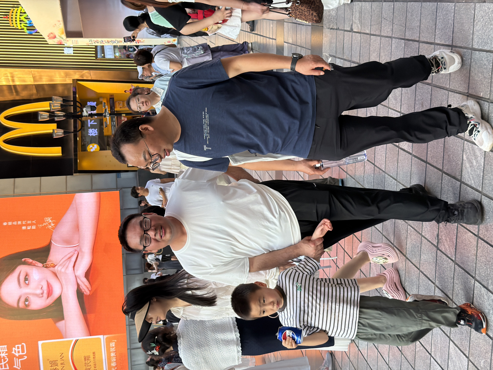 The image shows a street scene in Yuzhong District, China. In the foreground, two men and a young boy are walking together. The man on the left wears a white t-shirt and black pants, while the man on the right is in a navy blue t-shirt and black pants. The boy is wearing a striped shirt and grey pants and is holding a can of soda. A large digital billboard with an image of a woman is visible in the background, along with a McDonald's restaurant. The ground is paved with square tiles, and other pedestrians are visible in the background, suggesting a busy urban environment.