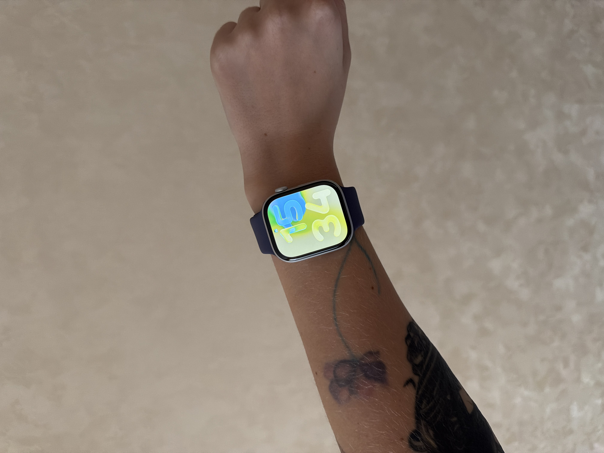 A person is wearing a silver Apple Watch with a dark blue band on their left wrist. The watch face displays a stylized green and blue digital clock reading 1:34. The person's arm is positioned diagonally in the frame. The forearm features a dark tattoo. The background is a textured beige wall. There is no indication that the image was taken in Kazan, Russia.