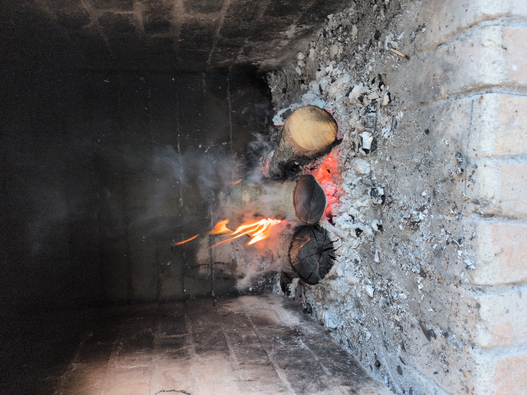 The image depicts a fireplace in Нежинка, Russia. Inside the brick fireplace, three logs are burning, with visible flames and smoke. Ashes surround the logs, covering parts of the brick structure. The left side of the fireplace is darker, contrasting with the ashy and brick-lined right side. There are no people visible in the image.