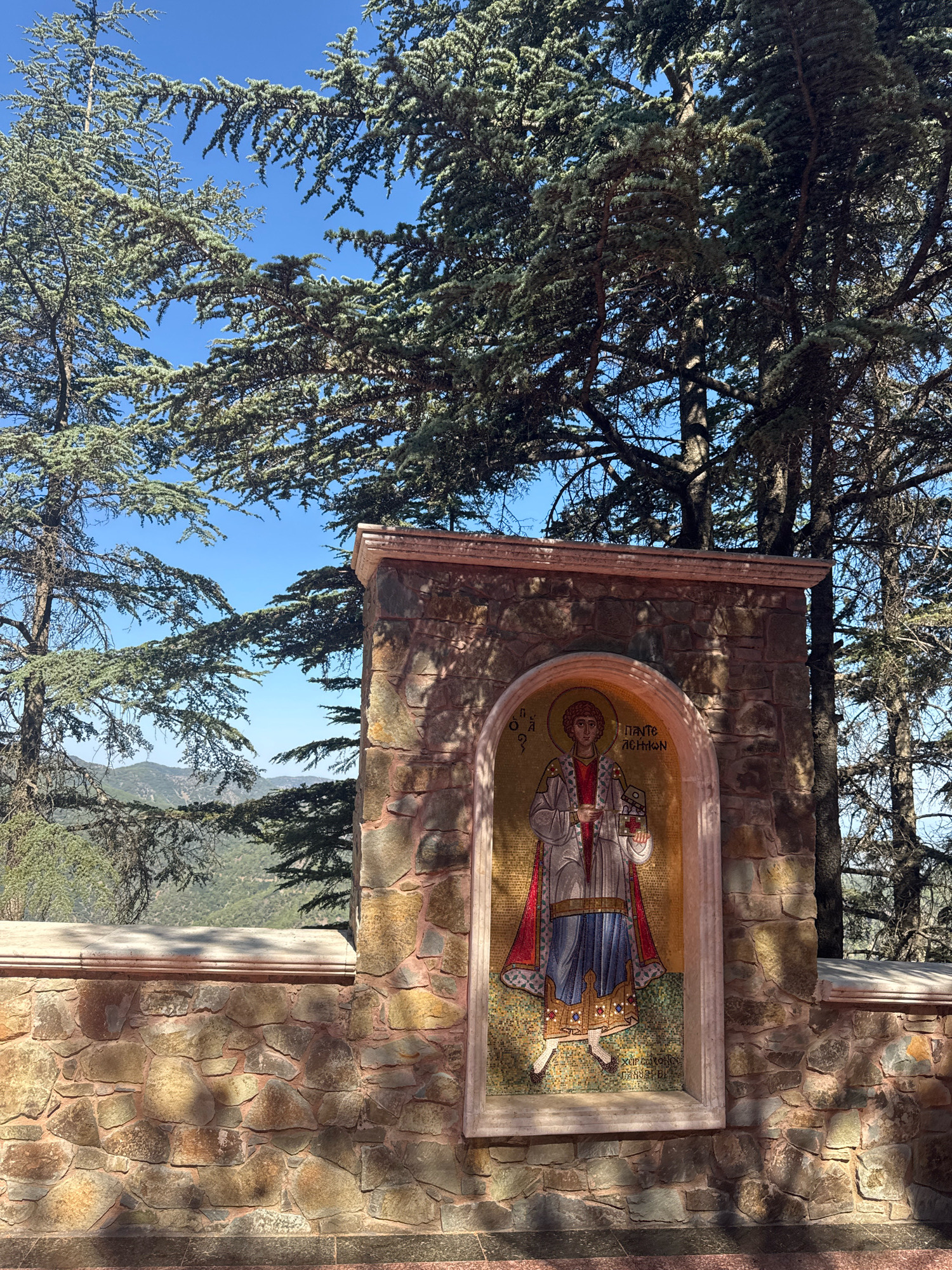 The image depicts an outdoor scene with a mosaic of a religious figure set into a stone wall. The mosaic is an arched panel featuring a figure with a halo, wearing traditional religious garments and holding what appears to be a book or sacred object. The figure is depicted in a mosaic style with intricate details and vibrant colors. The stone wall surrounding the mosaic is constructed from irregular, light-colored stones with darker grout. Above the mosaic, a decorative stone arch and lintel are visible. The wall is topped with a flat, lighter-colored stone ledge. In the background, tall evergreen trees with sparse foliage frame the scene, their branches reaching towards a clear blue sky. Beyond the trees, a lush, green mountainous landscape stretches into the distance, with some structures or a cross visible on a hilltop. The lighting suggests a sunny day, with dappled shadows cast by the trees on the wall and mosaic. This scene is located at the Monastery of Saint Nicholas of the Roof in Tsakistra, Cyprus, an area known for its natural beauty and religious significance.
