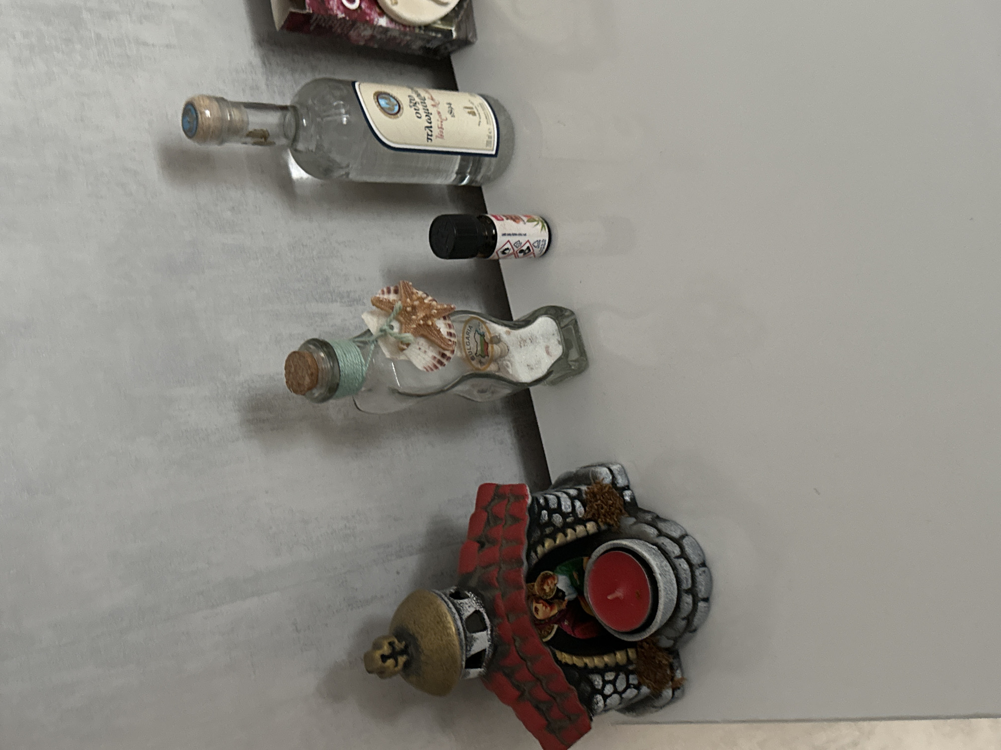 The image shows a collection of small decorative items attached to a grey surface, potentially a refrigerator door. From top to bottom, there's a bottle of Greek ouzo, a small vial with a black cap and a red and white label, a decorative bottle filled with sand and seashells, and a miniature decorative building with a red roof and a red candle inside. The location in the city of Lovech, Bulgaria is not apparent from the image.