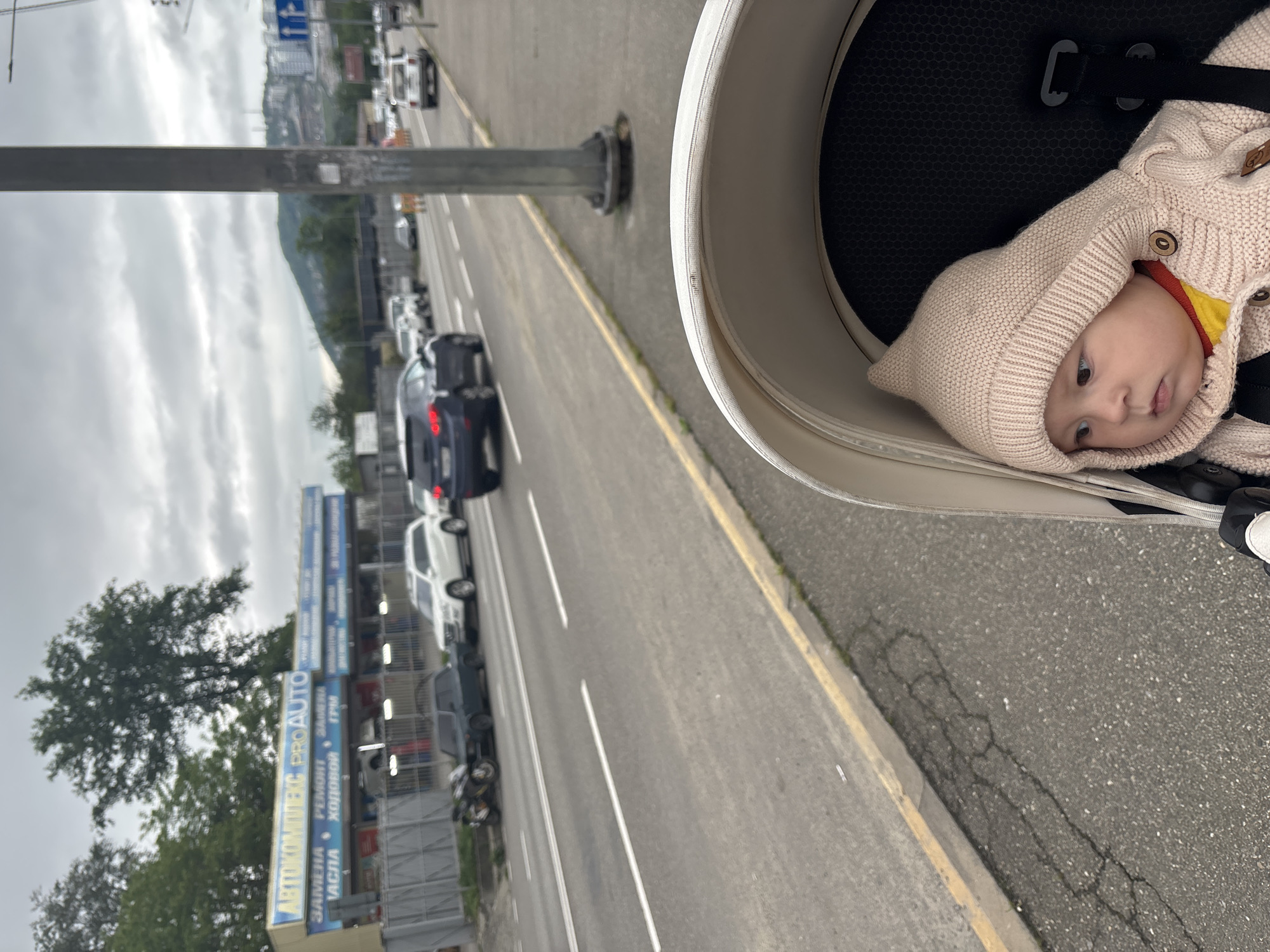 The image shows a baby wearing a beige hooded sweater in a stroller. The stroller is on the side of a road in Sochi, Russia. Cars are visible on the road, along with a utility pole. A building with "Автокомплекс PROAUTO" signage is present. Mountains are visible in the distance under a cloudy sky.