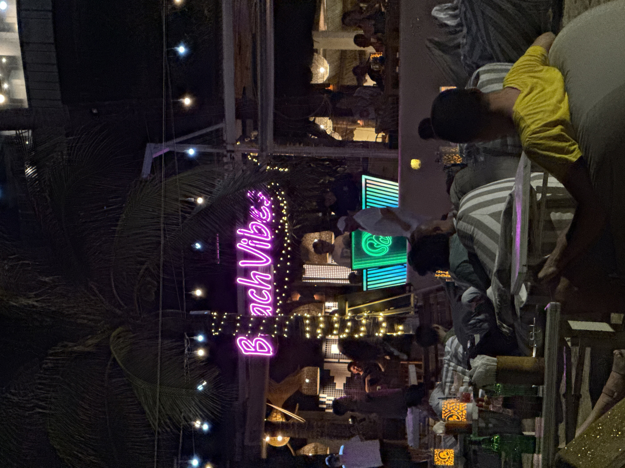The scene is an outdoor nighttime view in what appears to be a resort or beach club setting, likely in or near Nha Trang Ward, Vietnam, given the illuminated "Beach Vibes" sign in the frame. The neon sign is oriented vertically and is surrounded by palm trees, typical of tropical locations.
The foreground includes tables with people gathered, some interacting around food and drinks. There are candles and bottles on the tables, indicating an evening social event. Patrons are seated on striped cushions, with one individual wearing a yellow shirt and appearing to be leaning forward.
In the background, there are buildings with multiple floors and open structures where more people are gathered. There are also decorative lights strung overhead, contributing to the festive ambiance.
The image suggests a lively and casual atmosphere, likely at a beachside restaurant or lounge.