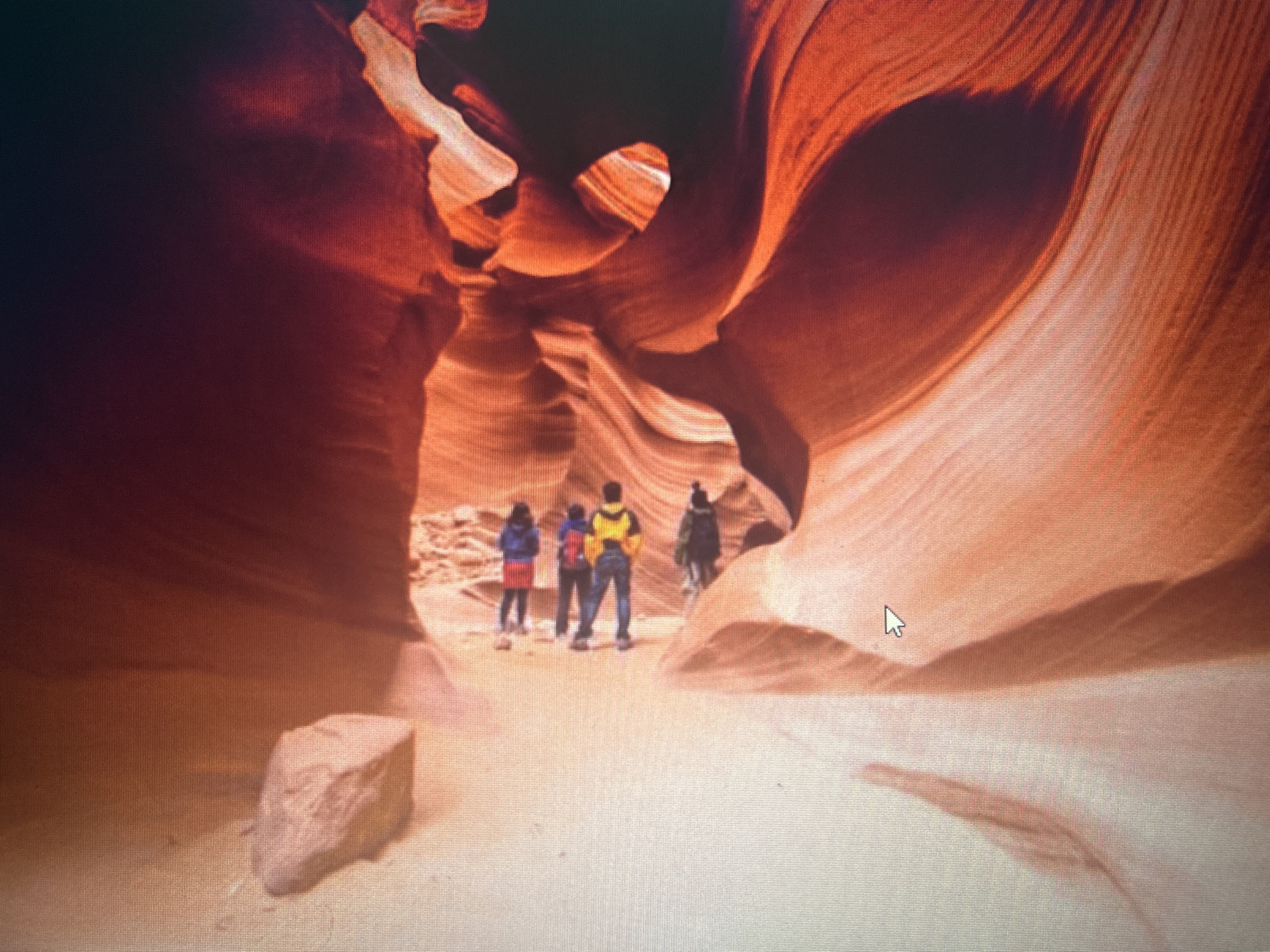 The image depicts a group of four individuals standing in a narrow canyon. The canyon walls are characterized by smooth, undulating rock formations in shades of orange and red, illuminated by natural light filtering from above. The ground is sandy and light-colored. The individuals, seen from behind, are dressed in casual outdoor clothing, including jackets and backpacks. The foreground features a large, rough-textured rock. The scene suggests exploration or tourism within a natural geological formation. There is no discernible connection to the city of Sofia, Bulgaria.