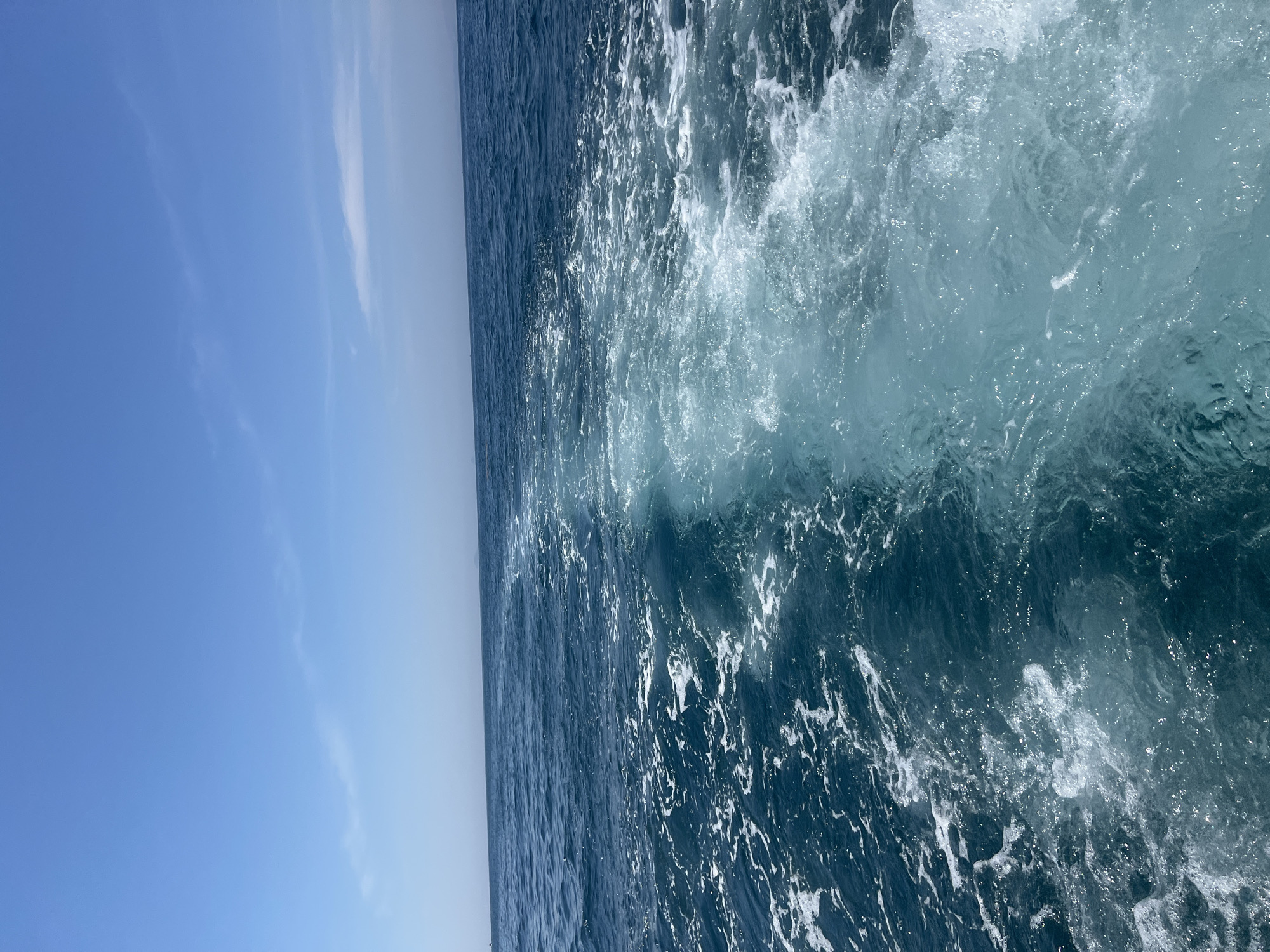 The image depicts a wide expanse of the ocean with a clear blue sky overhead. The foreground shows the turbulent wake of a boat, with frothy white water churning against the deeper blue of the sea. The horizon is visible in the distance, meeting the sky, and a faint landmass or island can be seen on the far right. The scene appears to be a daytime shot, likely taken from a moving vessel. Given the context provided, this scene is situated in the waters off the coast of Nha Trang, Vietnam, a popular coastal city known for its beaches and oceanic activities. There are no people or specific objects visible in the frame other than the natural elements of the sea and sky.