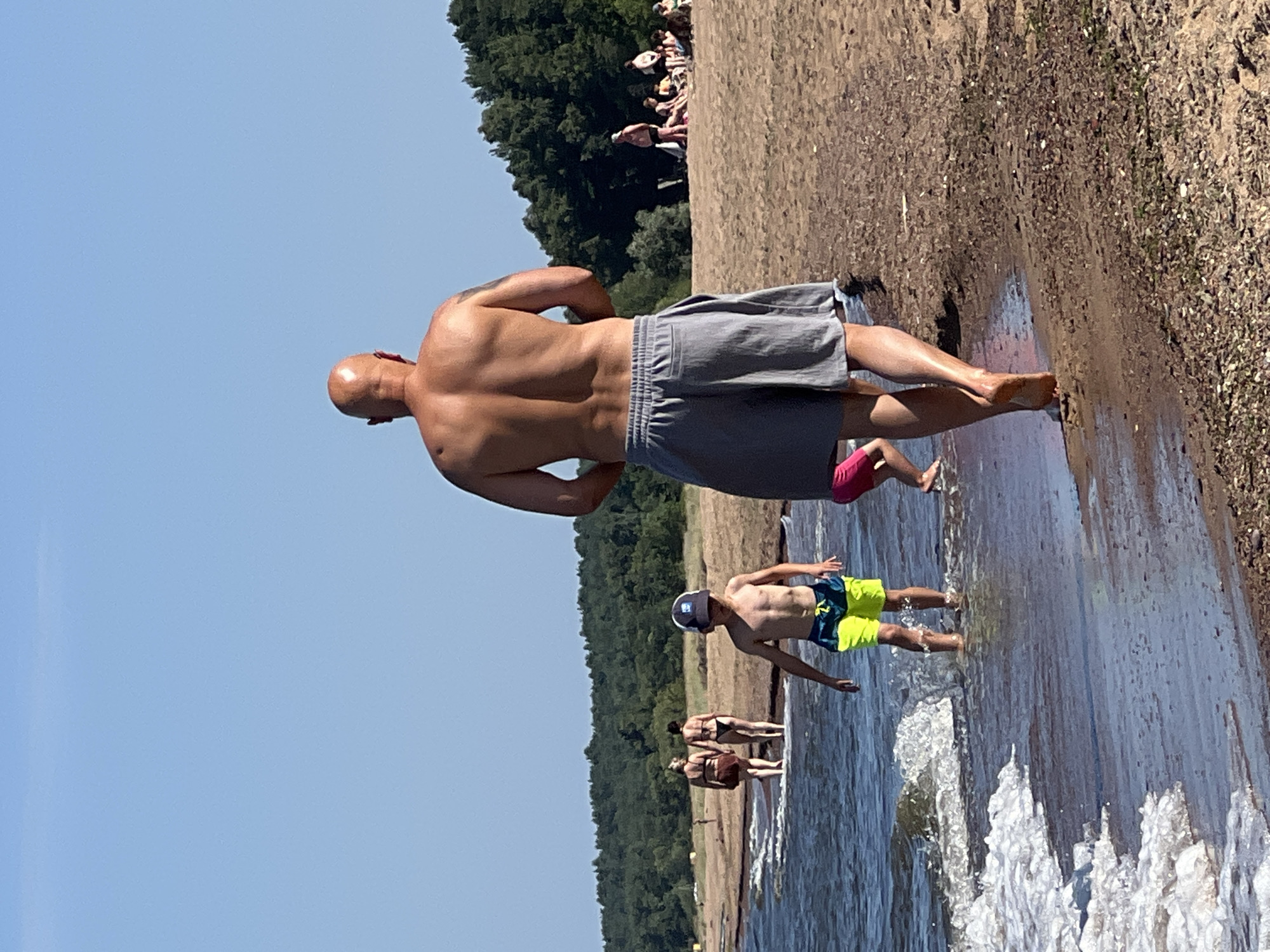 The image depicts a beach scene with several individuals enjoying the water and sand. In the foreground, a bald man wearing gray shorts walks away from the camera, his back to the viewer. To his left and slightly ahead, a young boy in neon green and blue swim trunks stands in the shallow water, facing away from the camera. Further in the background, a group of people are visible on the sand, some sitting and others standing. The water has small waves lapping at the shore, and the background is characterized by a line of green trees under a clear blue sky. The environment is a sandy beach with the sea.