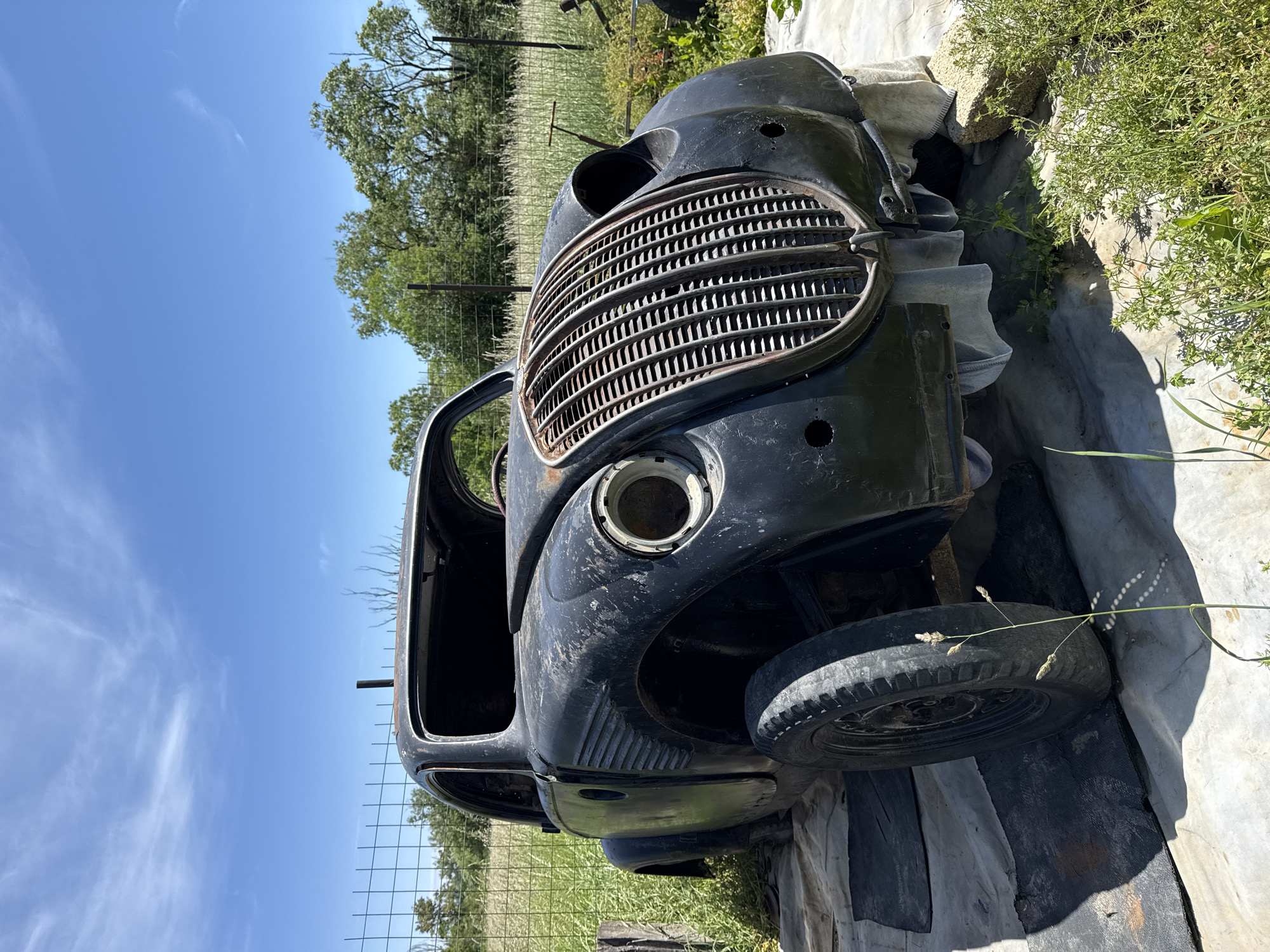 An old, dilapidated black car body sits outside in a rural environment. The car's grill is intact but rusted. The headlight casing on the driver's side is empty. One tire is visible. The car rests on a tarp or cloth. The background contains a blue sky, trees, and a wire fence. No people are present. The location is unspecified, although it is suggested to be in Omurtag, Bulgaria.