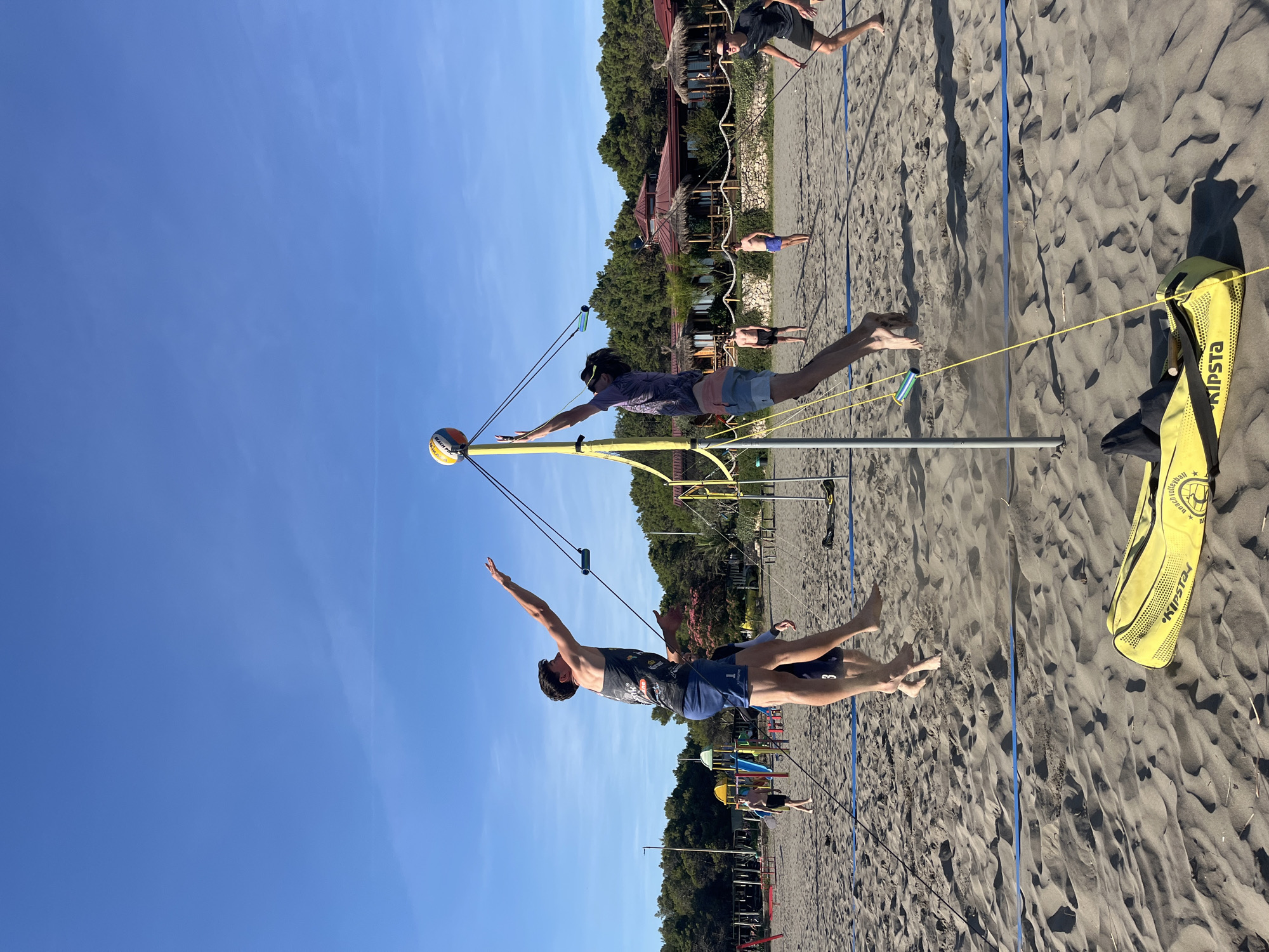 On a sunny day at the beach in Pistula, Montenegro, four young men are engaged in a game of beach volleyball. Two players are mid-air, jumping to hit the ball over the net, while two others are positioned to receive. The sandy court is marked with blue lines, and a yellow volleyball bag lies on the sand in the foreground. In the background, a line of trees and a restaurant with thatched roofs suggest a resort setting. The clear blue sky indicates fair weather.