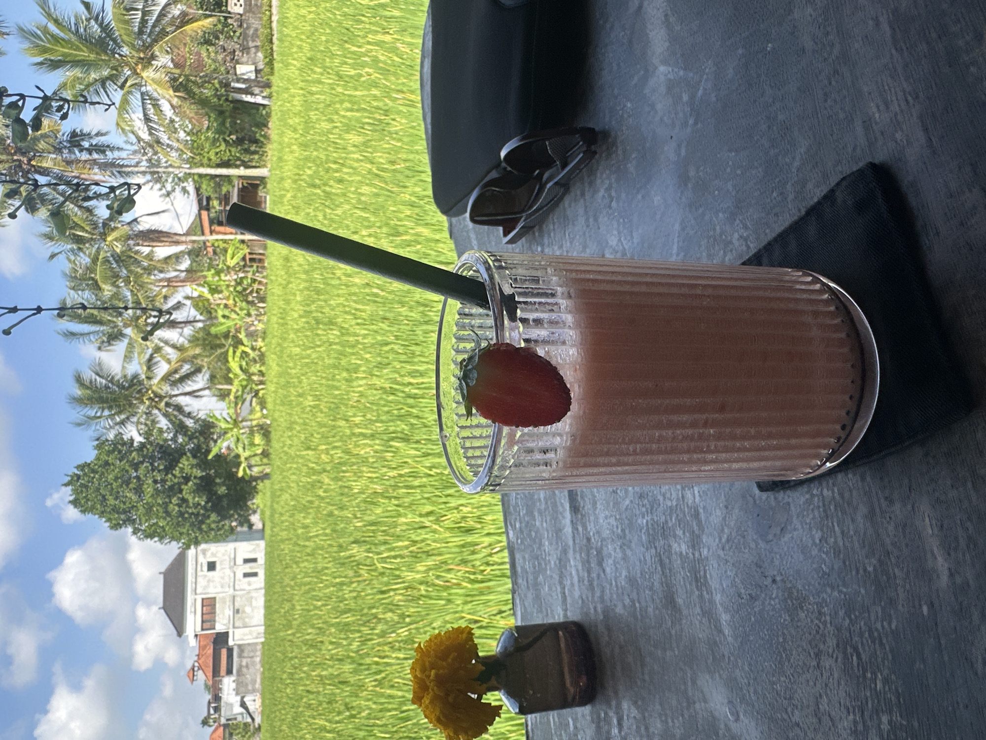 A close-up shot on a table shows a glass of pink smoothie garnished with a strawberry slice and a dark green straw. The glass is placed on a black coaster. To the left of the smoothie, a small brown vase holds a bright yellow flower. In the background, a lush green rice paddy stretches out under a partly cloudy blue sky, with palm trees and buildings dotting the horizon, suggesting a location in the Ubud District, Indonesia. A pair of sunglasses and a dark bag are also visible on the table.