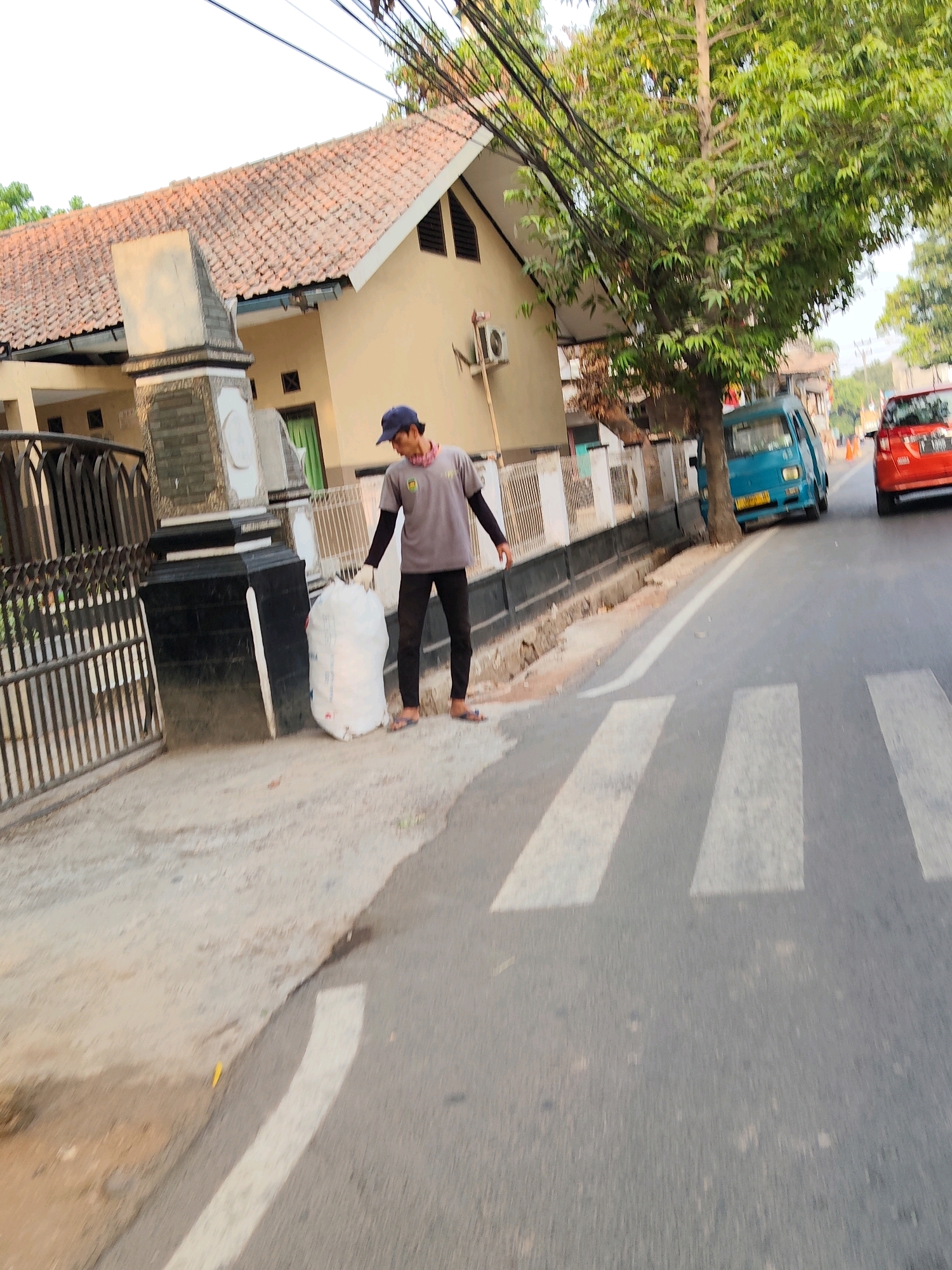 A man wearing a grey t-shirt, dark pants, a blue cap, and a red bandana around his neck stands on the side of a street in Purwakarta, Indonesia, holding a large white garbage bag. He is positioned near a zebra crossing and a fence in front of a building with a tiled roof. A blue van and a red car are visible on the road, with trees and power lines overhead. The scene appears to be a residential or commercial area with sidewalks and street infrastructure.