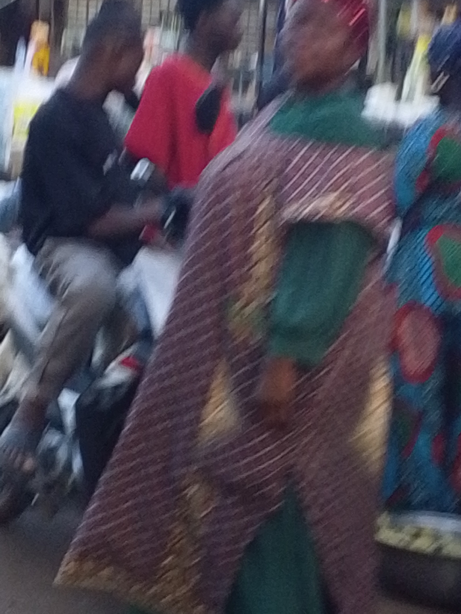 The image shows a street scene in Tamale, Ghana. In the background, there are two men, one wearing a black shirt and light-colored pants, sitting on a motorbike. The other man is wearing a red shirt. In the foreground, there are two women. The woman in the center is wearing a green dress and a patterned outer garment with horizontal stripes. The woman to her right is wearing a blue patterned dress. The scene appears to be a crowded market or street with people passing by.