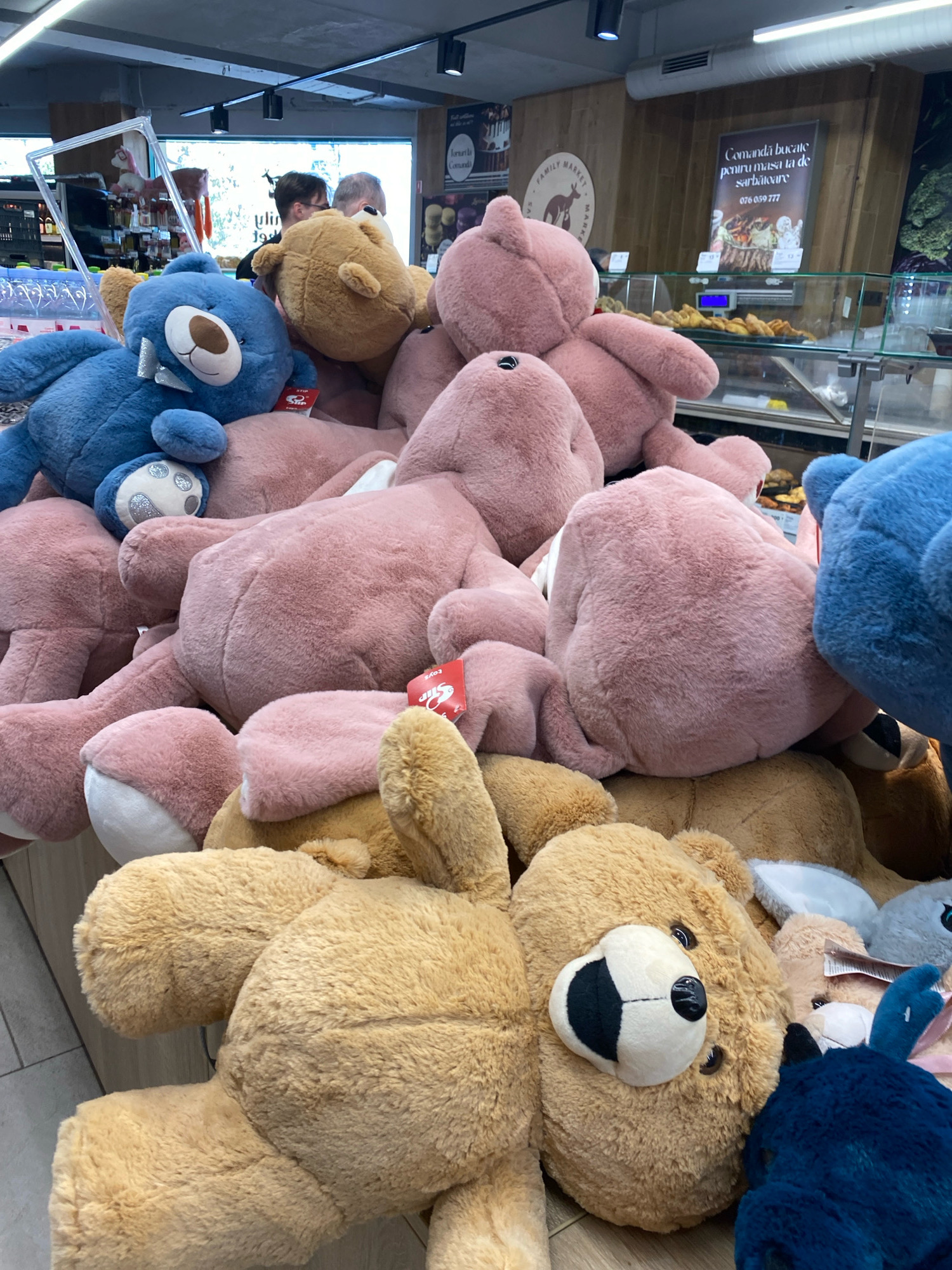 The image depicts a retail setting, likely a supermarket or a market stall, with an abundance of plush teddy bears of various sizes and colors, predominantly pink and tan, with a few blue ones visible. The teddy bears are piled on top of each other, filling the foreground and extending into the middle ground. In the background, a counter displays food items, suggesting a deli or bakery section. A sign with Romanian text, "Comanda bucate pentru masa la de sărbătoare" and a phone number, is visible on the wall behind the counter. Two individuals are partially visible in the background, one wearing glasses and looking towards the right. The lighting appears to be artificial. The location is identified as Chișinău, Moldova, implying this is a market or store within that city.