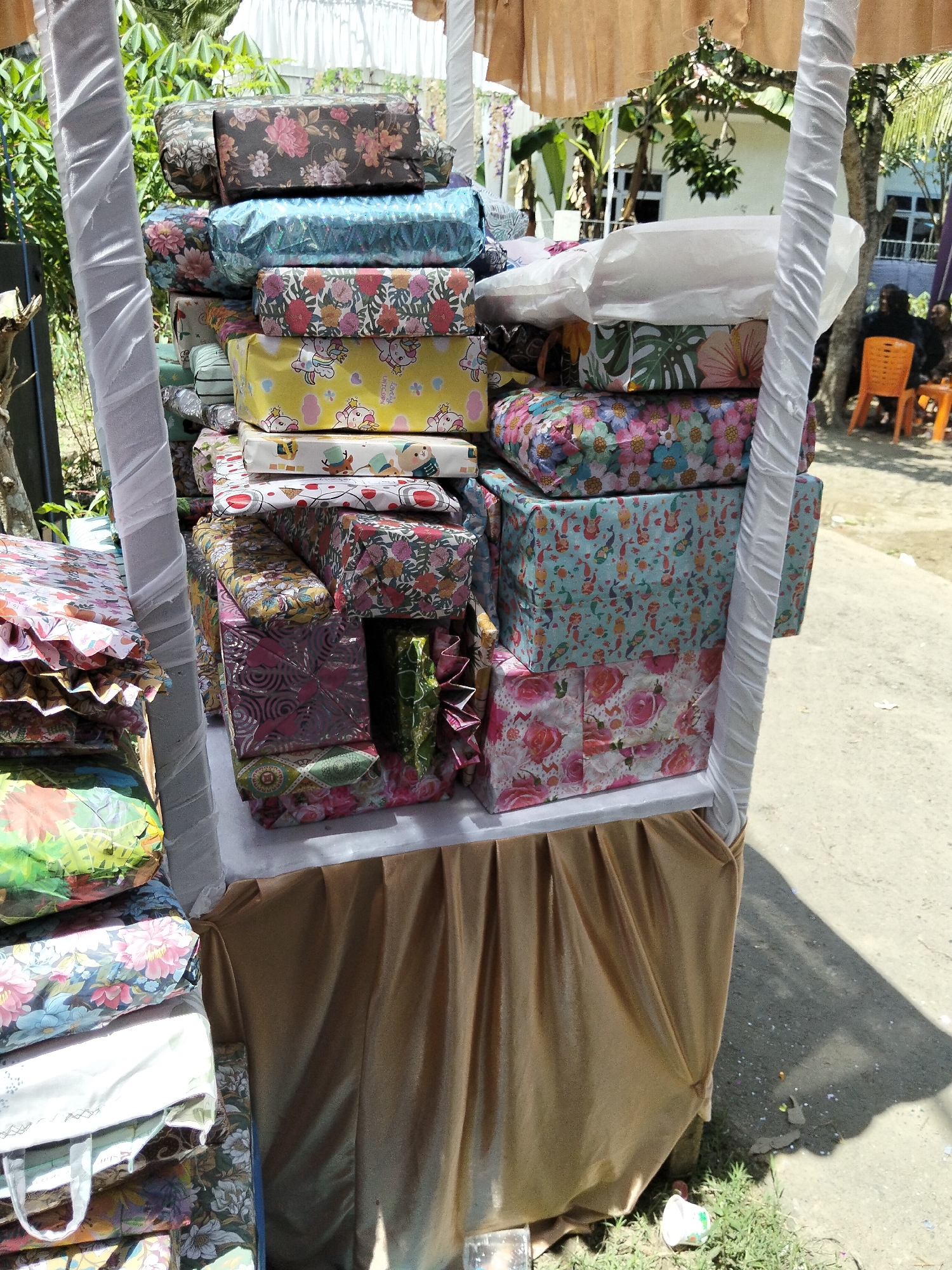 In the city of Pulonga, Indonesia, a stall display features a tall stack of wrapped presents. The presents are of various sizes and wrapped in colorful paper with floral and cartoon patterns. The stall itself is draped in a tan-colored fabric. In the background, trees and a building are visible. To the right, two individuals are seated on orange chairs. The ground around the stall is a dirt path.