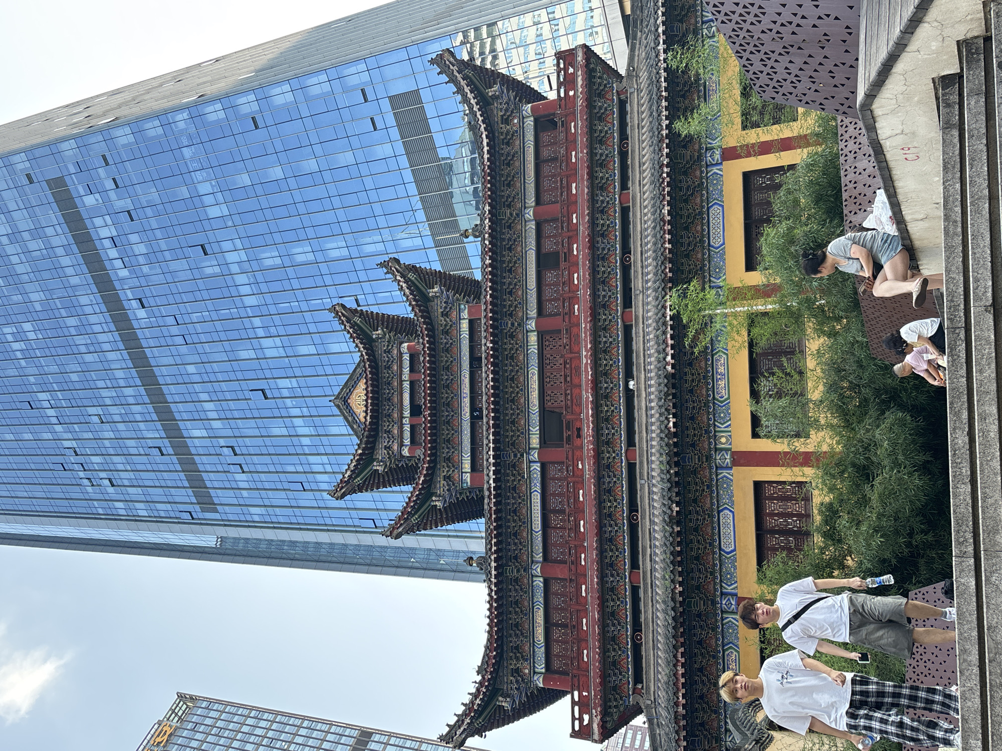 A vertical, wide shot captures a juxtaposition of modern and traditional architecture in Yuzhong District, China. Dominating the left half of the frame is a towering, contemporary glass skyscraper, its blue panes reflecting the sky and the traditional building. In the center and right, an intricately detailed, multi-tiered traditional Chinese building stands, adorned with red, gold, and patterned decorations and upturned eaves. Lush green trees provide a natural element, interspersed between the two architectural styles.
Several people are visible in the scene. In the foreground, two young men are standing. The one closer to the camera is wearing a white t-shirt and plaid pants, holding a water bottle. The other, slightly further back, wears a white t-shirt, gray shorts, and a black strap across his chest, also holding a water bottle. Further up and to the right, a woman in a gray shirt and denim shorts is seen kneeling or bending down, with another person, possibly a child, visible to her right.
The time of day appears to be daytime, with bright lighting suggesting clear weather, though some clouds are present in the sky. The scene seems to be in an urban setting, likely a public area where modern development meets preserved cultural heritage. The overall impression is one of contrast and harmony between different eras and styles. There is some faint, illegible red graffiti on the concrete structure on the far right.
