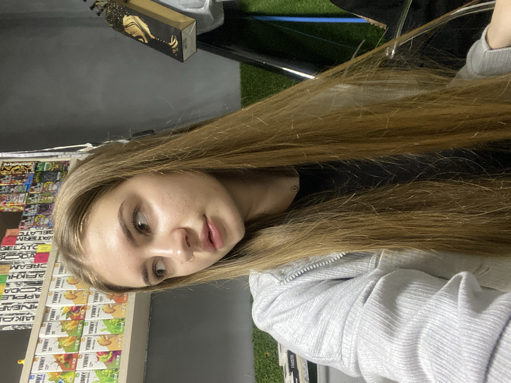 The image depicts a young woman with long, light brown hair, seen from the chest up, looking towards the right side of the frame. She wears a light grey, ribbed, zip-up hoodie over a dark-colored garment, possibly black. A small silver nose ring is visible on her left nostril, and a delicate necklace with a small 'D' pendant adorns her neck.
The background reveals an indoor retail environment, strongly indicative of a vape shop. To the left, a white shelf displays numerous small, rectangular boxes primarily branded "YUMMY," featuring various fruit graphics (e.g., apple, peach, grape, pineapple). Other vape liquid or product brands like "LAIKA," "APPLE SODA," "ARKHI CREAM," and "WATERM GELATO" are visible on an upper shelf. A small yellow duck figurine sits on the top shelf.
To the right, a plain dark grey wall serves as a backdrop. A black object with a gold, stylized design, possibly a "CROWN" branded vape product box, hangs against the wall. Below this, a section of green artificial turf is visible. Another white item with "FAVOSTIX" text is partially visible on the bottom left, positioned on a surface with the artificial turf behind it. A dark, possibly conical or cylindrical object stands vertically against the turf on the far right.
Regarding the specified location, no visual information within the image confirms the specific geographic location as the city of Дьяково, Russia, or any other specific city or country. The observed Cyrillic text on product packaging is consistent with Russian-speaking markets, but not specific to a single city.