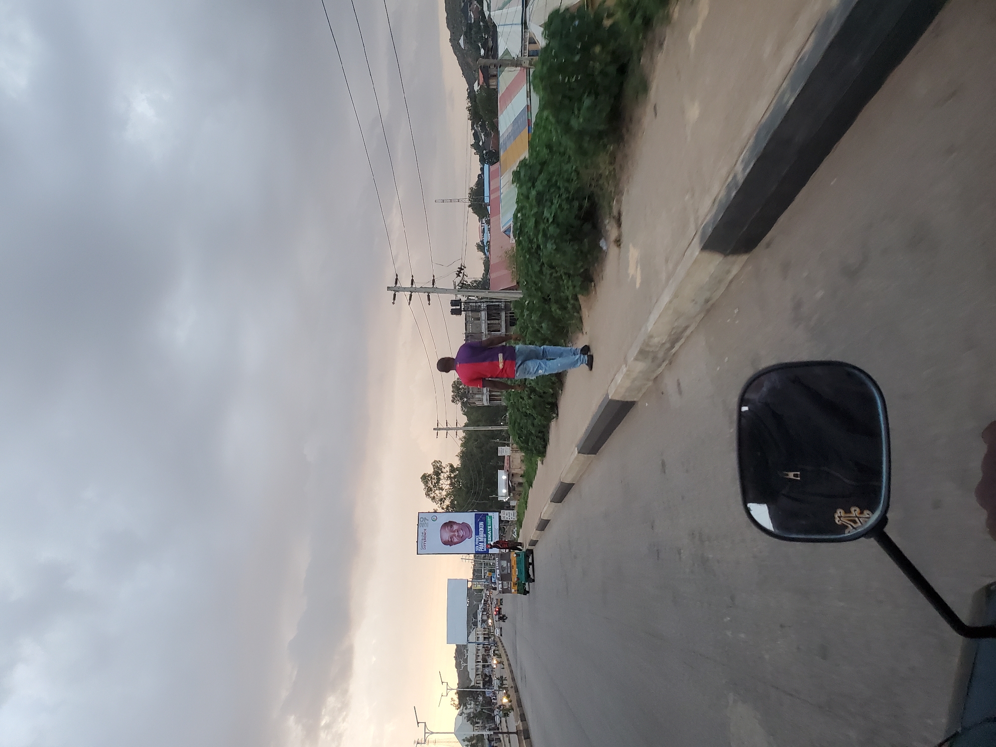 The image captures a street scene in Gura Top, Nigeria, with an overcast sky. In the foreground, the perspective is from a motorcycle, with its side mirror visible on the left side, reflecting a person wearing a dark jacket. A man wearing a purple and red t-shirt and blue jeans is walking away from the camera on the right side of the road, along the sidewalk. The road itself is paved and appears to be a main thoroughfare, with vehicles and a tuk-tuk visible in the distance. On the left side of the road, there is a billboard featuring a portrait of a man and some text. The environment includes buildings with colorful roofs and lush green vegetation lining the sidewalk. The overall impression is of a busy urban environment under a cloudy sky.