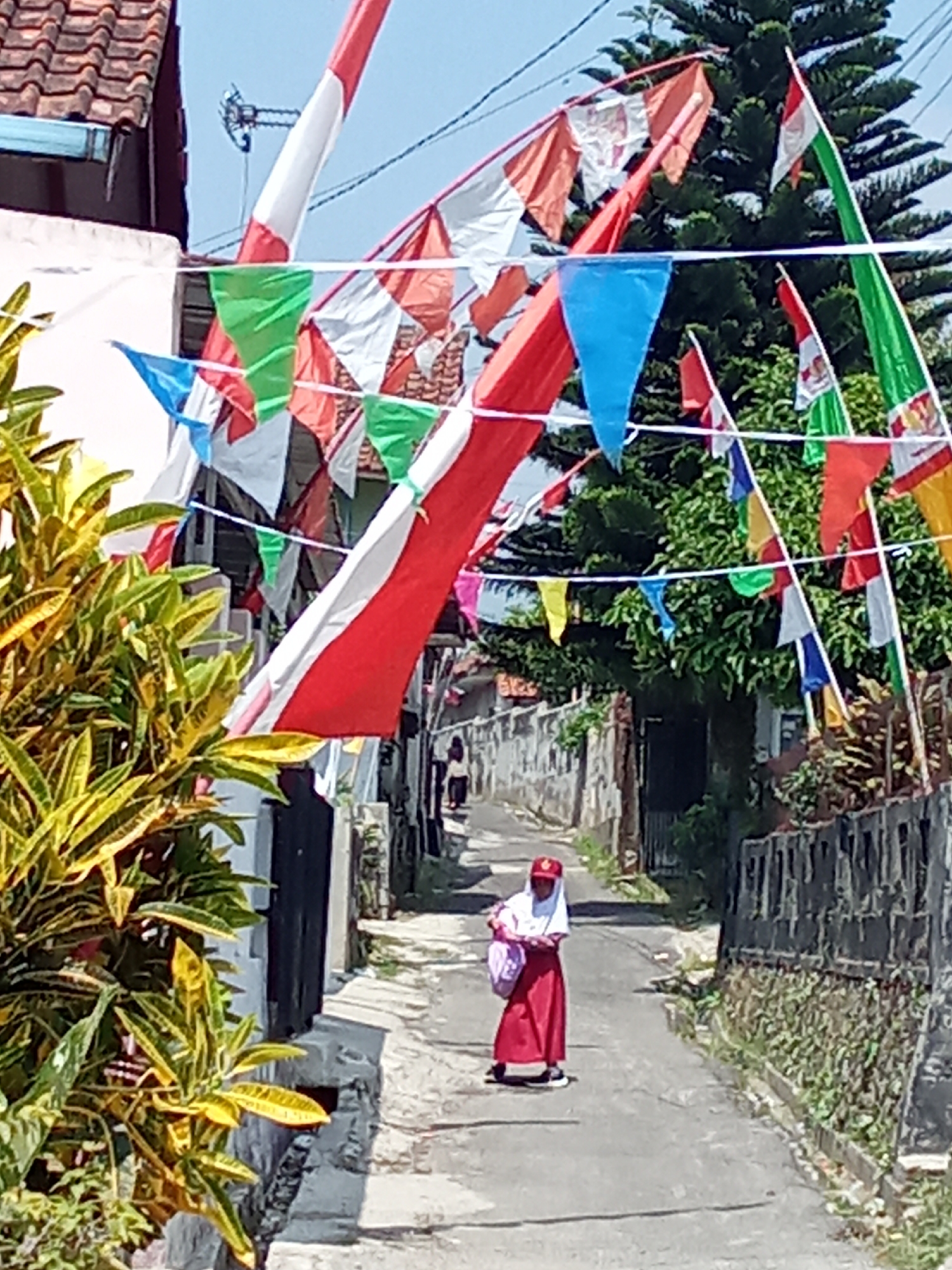 The image depicts a street scene in the city of Subang, Indonesia, decorated with numerous colorful flags and bunting, likely for a celebration. The flags, primarily in red and white, are strung across the narrow street, creating a festive atmosphere. In the foreground, lush green and yellow foliage is visible on the left. The street itself is paved and appears to be a residential area with houses lining both sides.
A young student, dressed in a red skirt and white hijab, is the central human subject. She is walking away from the camera, carrying a light pink bag over her shoulder. Her stance suggests she is mid-stride. In the background, another person is visible further down the street, walking in the same direction. The architecture of the houses, with their tiled roofs and simple facades, is characteristic of Indonesian residential areas. The overall environment is bright and sunny, indicating a clear day.
