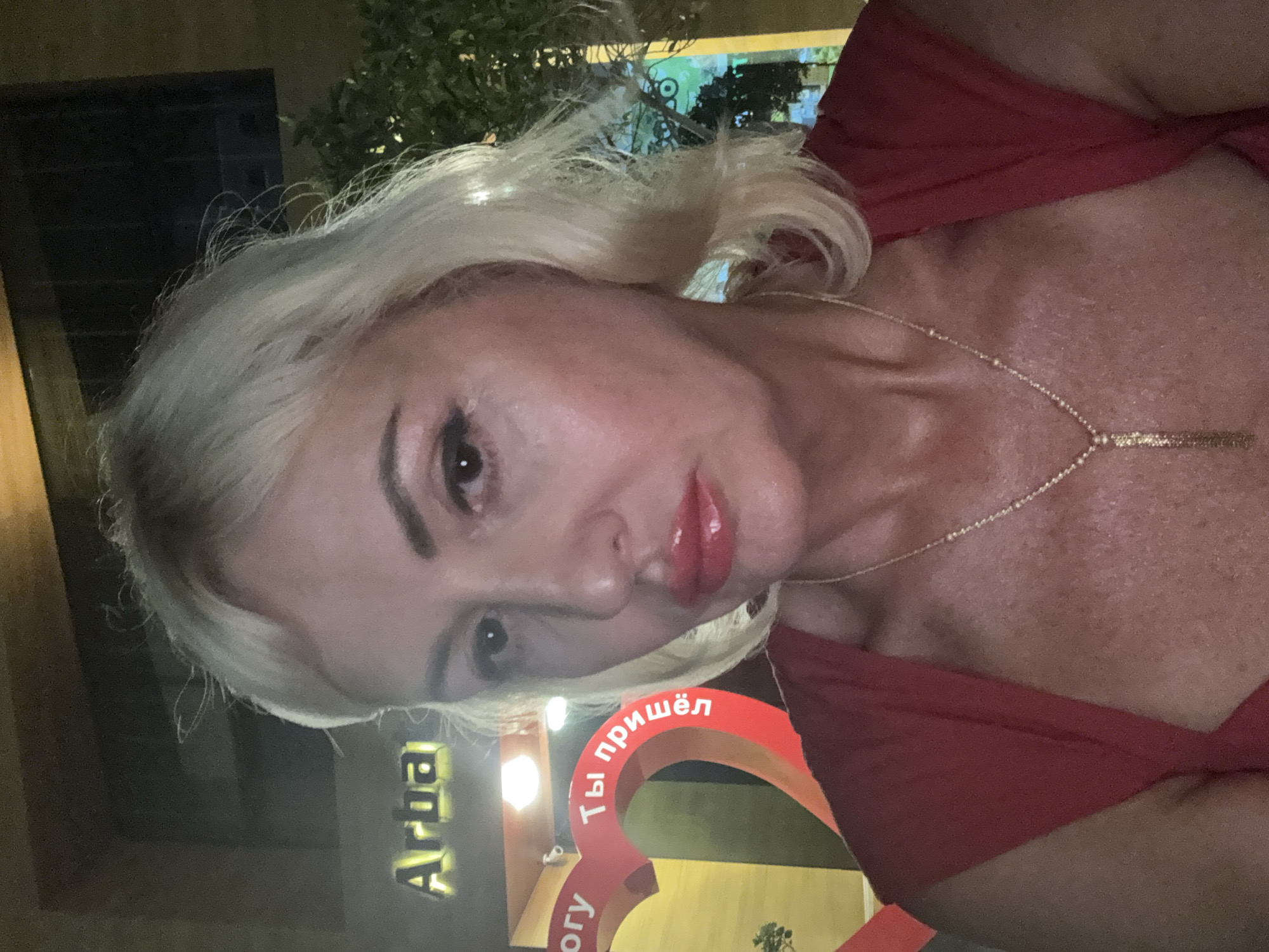 The image shows a close-up, selfie-style portrait of a middle-aged Caucasian woman with blonde hair, heavy makeup including bright red lipstick, and a red dress. She is wearing a gold necklace with a pendant. The background is blurred and appears to be outdoors at night, possibly a street or an establishment. There is a sign in the background that reads "Arba" in yellow illuminated lettering. To the left of the woman, there is a large, red, heart-shaped decoration with text in white. Part of the text on the heart is visible and appears to be in Russian, with the words "Ты пришёл" (You came) being legible. The overall lighting suggests it is evening or nighttime. The context of the "Arba" sign and the Russian text on the heart suggest a potential location in Alanya, Turkey, which has a significant Russian-speaking tourist population and establishments catering to them.