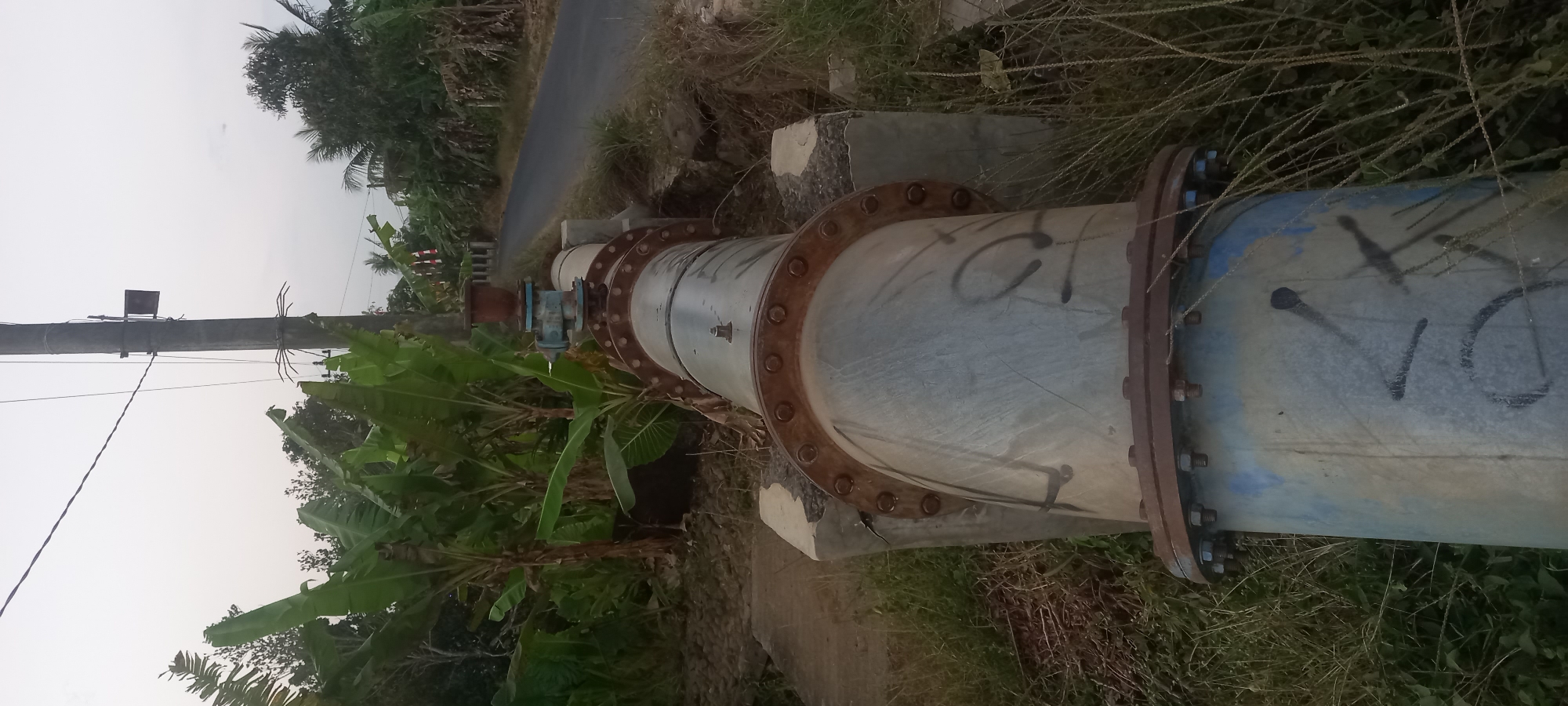 A large, grey pipe with some blue and black graffiti runs diagonally across the frame, supported by concrete structures. The pipe has rusted flange connections with numerous bolts. A valve is visible on top of the pipe. The scene is outdoors, with lush green banana trees to the left and right. A paved road is visible in the background, with a utility pole standing nearby. The sky is overcast. The image appears to be taken in Sidamulya, Indonesia, given the presence of banana trees and the general environment. No people are present in the image.