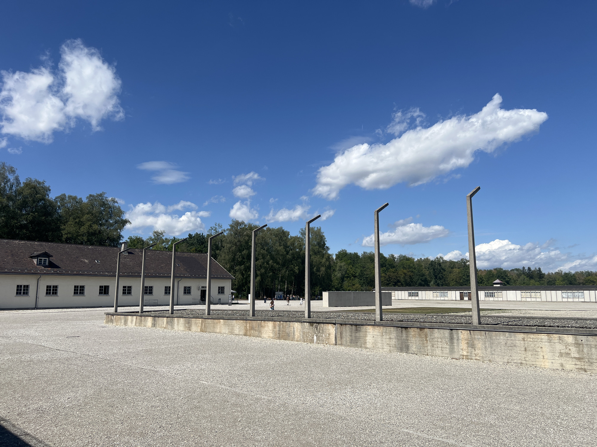 The image depicts an outdoor scene at the Dachau Concentration Camp Memorial Site in Dachau, Germany. In the foreground, there is a large, gravel-covered area with a concrete retaining wall. Several tall, slender metal poles, presumably remnants of a former structure, are visible in the middle ground. Beyond the retaining wall, there are several long, white buildings with dark roofs and numerous windows. These buildings are situated on a gravel-covered ground. In the distance, a line of trees forms the background, with a bright blue sky and scattered white clouds above. Several people can be seen walking in the middle ground, between the buildings and the poles. The overall atmosphere appears somber and reflective.