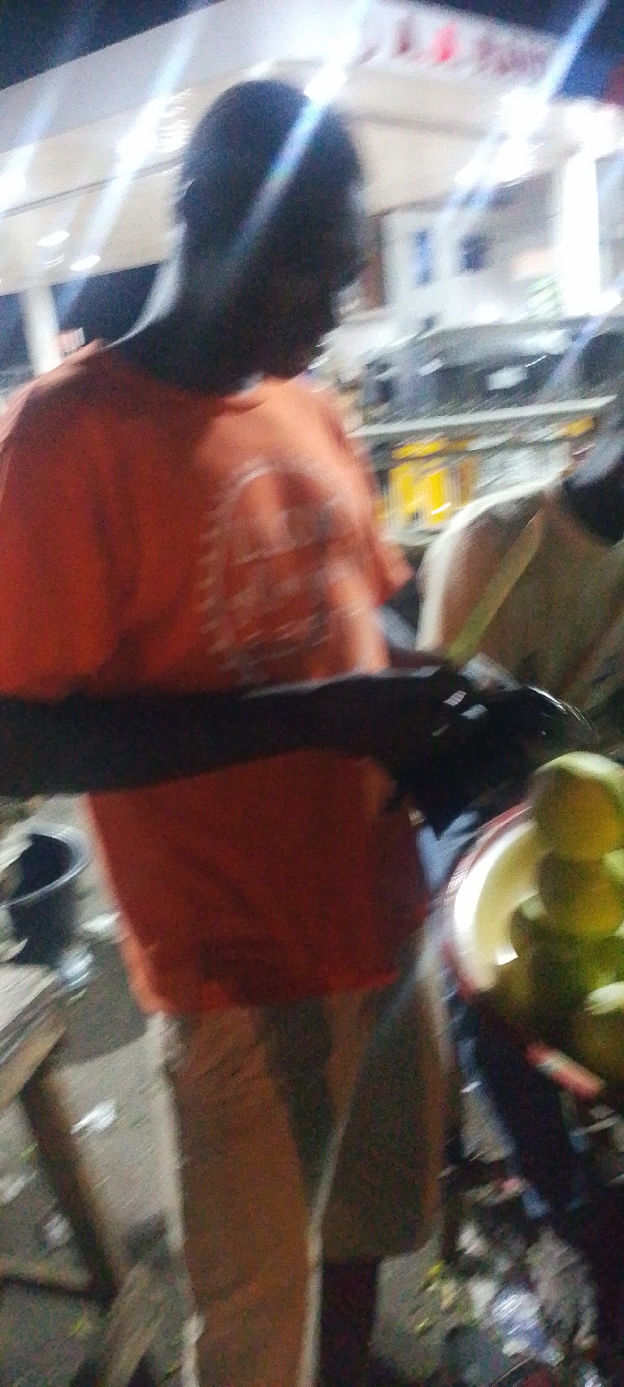 In Lokoja, Nigeria, an individual wearing an orange T-shirt and light-colored shorts is shown preparing fruit, possibly mangoes, with a knife near a street-side setup. Another person, wearing a light shirt, is partially visible to the right. There is a bucket and some objects on a wooden structure in the background. Street clutter is visible in the lower part of the frame, with a building lit by overhead lighting visible in the background.
