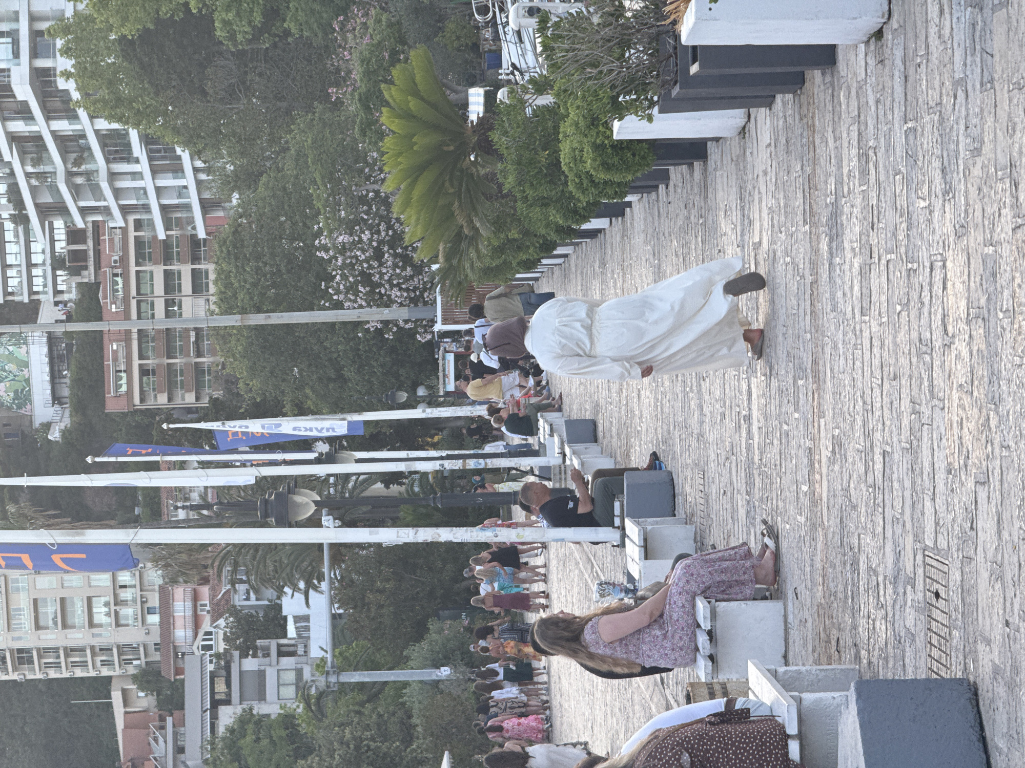 This image depicts a pedestrian promenade in Budva, Montenegro, likely during late afternoon or early evening. The promenade surface is paved with light-colored, irregular stone blocks.
In the foreground and midground, several individuals are present. On the left, a woman in a floral-patterned dress sits on a white concrete bench, facing right. Her feet, wearing sandals, are visible. Another person is partially visible to her left on an adjacent bench. Further along the left side of the promenade, a man in a dark t-shirt and light trousers sits on another white bench, appearing to interact with a handheld device. White, vertical poles with attached lamps and blue flags featuring orange symbols and text (e.g., "MAKS") line the left side, extending into the background.
On the right side of the promenade, a person wearing a long, loose-fitting white dress and sandals walks away from the camera. This side of the pathway is bordered by a series of dark and white planters containing green shrubs and small trees, culminating in a large sago palm.
In the mid-distance on the left, a group of people, primarily women in various dresses, are walking and standing, suggesting a social gathering or stroll. Other individuals are scattered further along the promenade.
The background features multiple multi-story buildings, including modern structures with balconies and older buildings with reddish roofs, situated on green, tree-covered hillsides. Various trees, including flowering ones with pink and white blossoms, are visible throughout the scene, flanking the promenade and covering the hills. A drainage grate is present in the promenade's paving near the lower left. Elements suggestive of a marina or waterfront are visible on the far right edge of the image.
