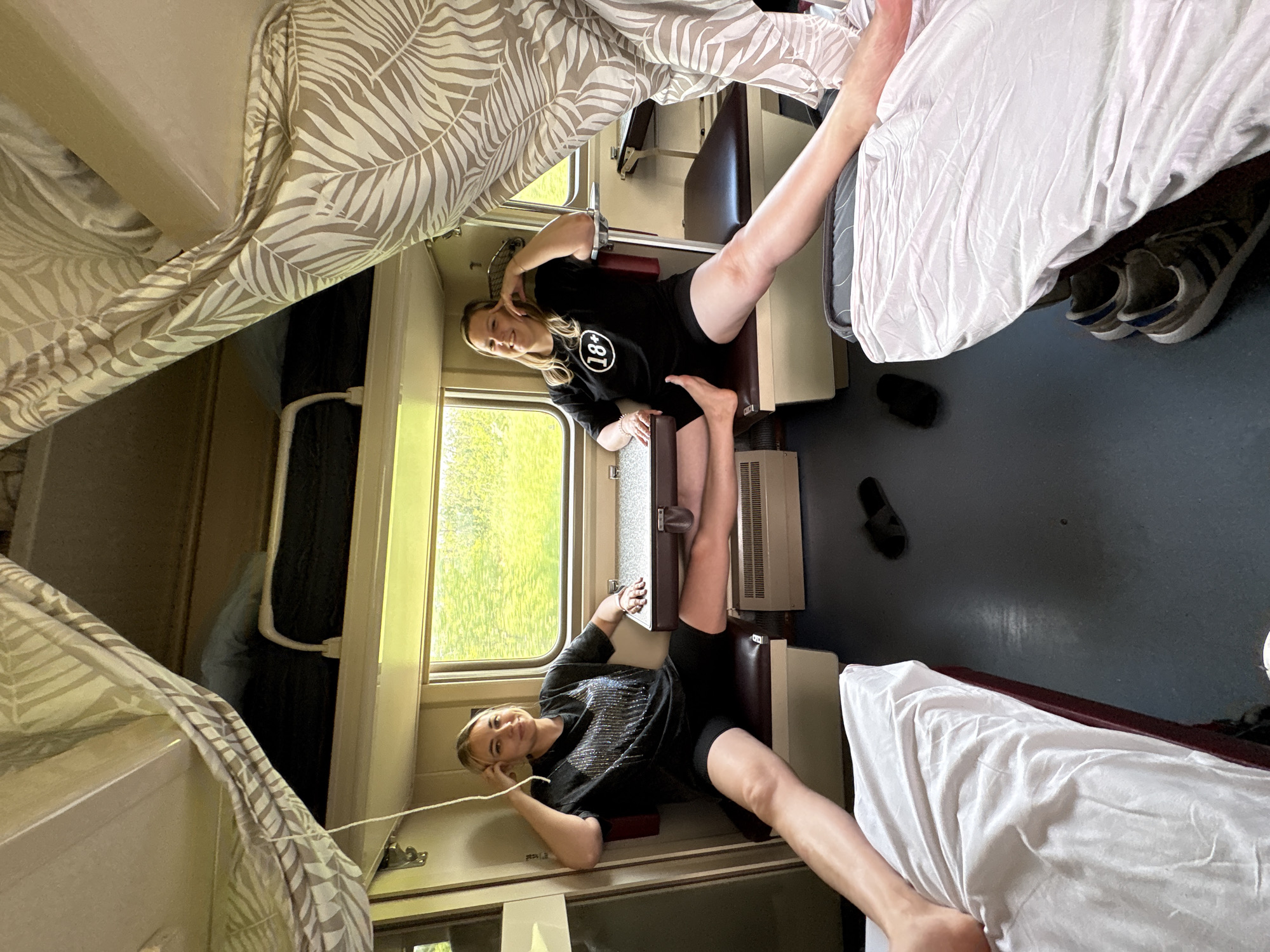The image shows the interior of a train compartment. Two women are seated on lower bunks. One woman is wearing a black t-shirt with a large "18+" logo. Bedding is visible on the upper bunks. Luggage is stored near the upper bunks. Shoes and sandals are on the floor. The scene is not in Eremeevka, Russia. It is in a train car.