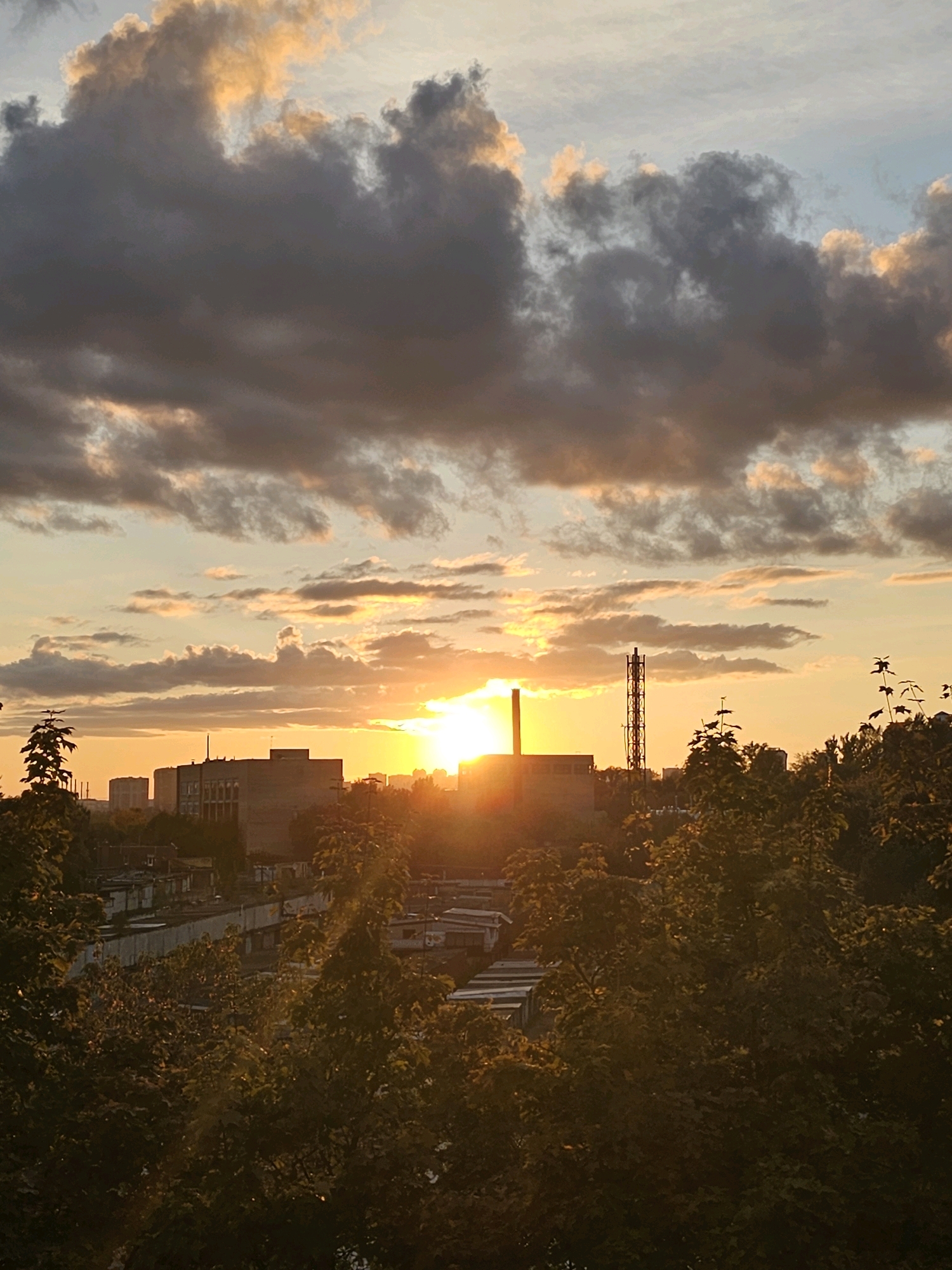 The image depicts a sunset over the city of Khimki, Russia, likely in the early evening. The sky is filled with dramatic clouds, ranging from dark grey to tinged with orange and yellow from the setting sun. The sun itself is a bright, intense orange disc, partially obscured by clouds, casting a warm glow across the scene. In the midground, industrial buildings and structures are visible, including a tall, lattice-like tower that appears to be a communication or transmission tower. Below the industrial elements, there are rows of smaller buildings and what seem to be garages or storage units. In the foreground and on the right side of the image, dense green trees, partially illuminated by the sunset, frame the view of the city. The overall impression is one of an industrial urban landscape bathed in the warm, fading light of dusk. There are no people or animals visible in the image.