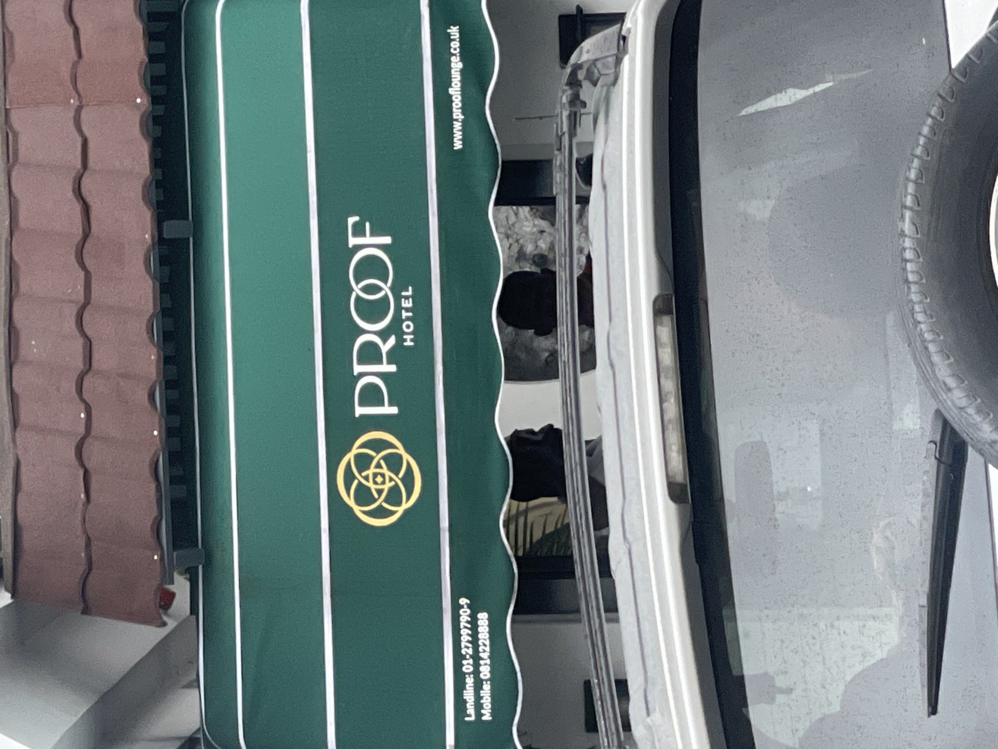 The image shows the exterior of a hotel named "PROOF HOTEL" located in Ilado, Nigeria. The hotel has a green awning with white lettering and a logo. Below the awning, there is a windshield of a vehicle, partially obscuring the view of the hotel entrance. Two individuals are visible as silhouettes through the windshield, appearing to be seated inside the vehicle. The overall scene suggests an urban environment with a focus on the hotel's branding and its immediate surroundings.