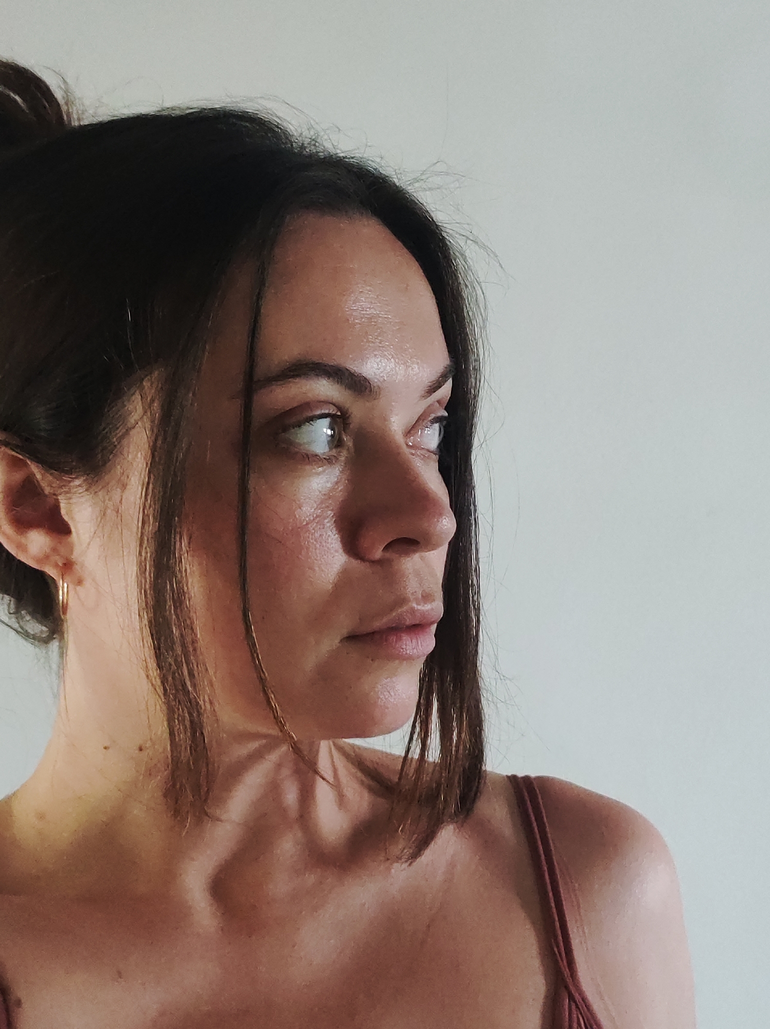 A head-and-shoulders portrait of a light-skinned woman with dark brown hair pulled back in a bun with a few strands framing her face. She is facing to the right, looking off-camera with a pensive expression. She wears a thin gold hoop earring in her left ear and a reddish-brown spaghetti-strap top. The background is a plain, off-white wall. The city and precise location are unknown.