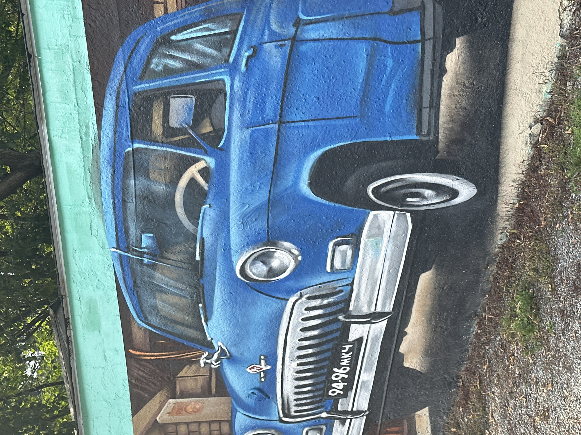 The image is a photograph of a mural painted on a brick wall, depicting a blue vintage car. The car is shown from the front, slightly to the left, with its grille, headlights, and a portion of the hood visible. A license plate with the characters "94-96МКЧ" is prominently displayed on the front bumper. The car has a chrome grille, a hood ornament of a leaping animal, and a red emblem. The mural is rendered with a textured, almost three-dimensional effect, giving the impression of a real car. Above the mural, a teal-colored overhang and green foliage are visible, suggesting an outdoor setting. The bottom of the image shows a gravel path and some dry grass. The overall scene is a vibrant street art display.
