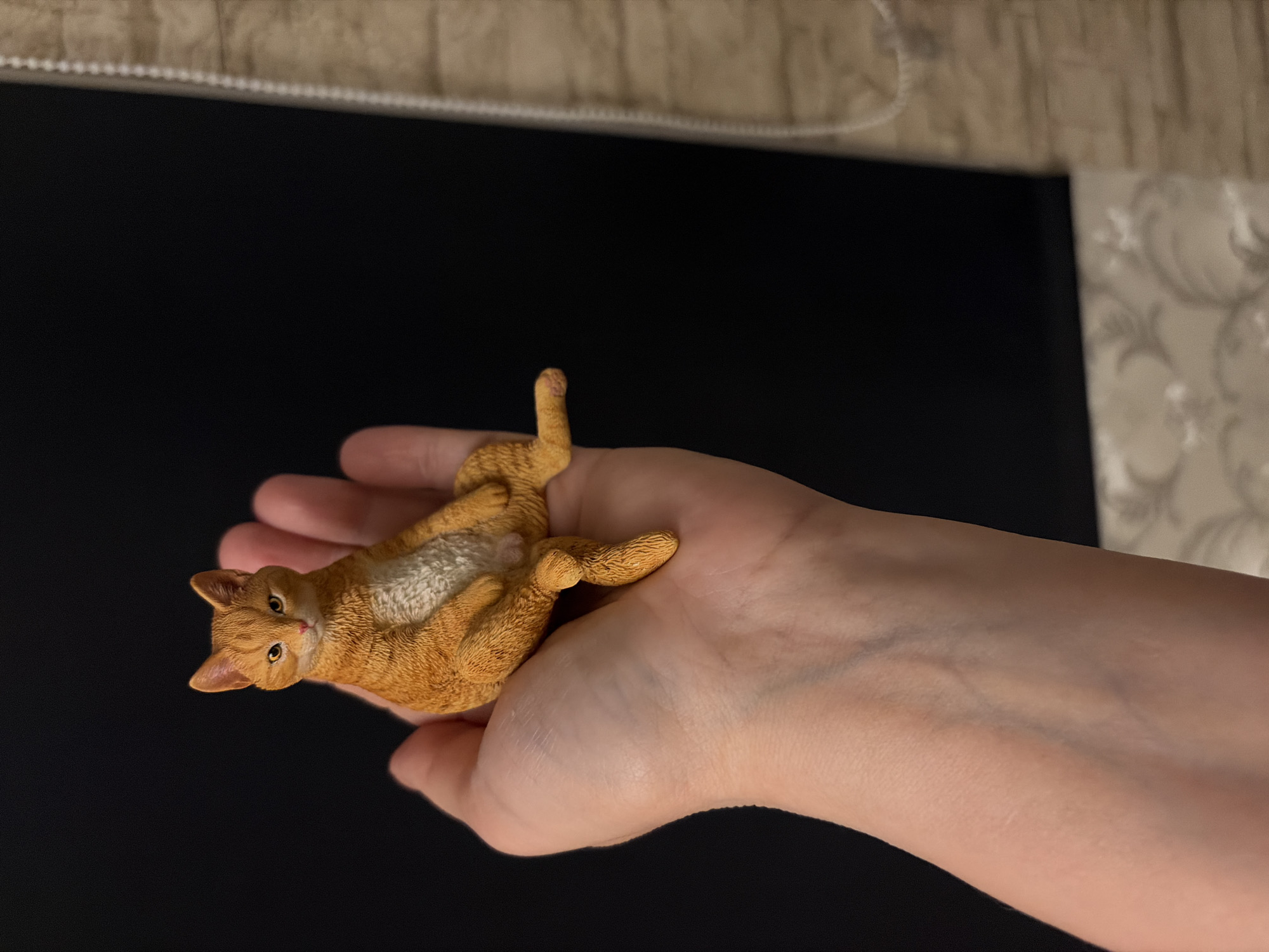 A small, orange, resin cat figurine is held in the palm of a person's hand. The cat is sitting with its legs crossed and its body tilted slightly to the left. The background is divided into two sections: the left side is a solid black color, and the right side shows a textured, beige wall with a white cord hanging vertically. The cat's fur is detailed, and its eyes are a piercing yellow. The hand holding the cat is fair-skinned and appears to be resting on a surface that is out of focus. The lighting in the scene is dim, creating shadows on the cat and hand. The scene does not appear to be located in the city of Härryda, Sweden.