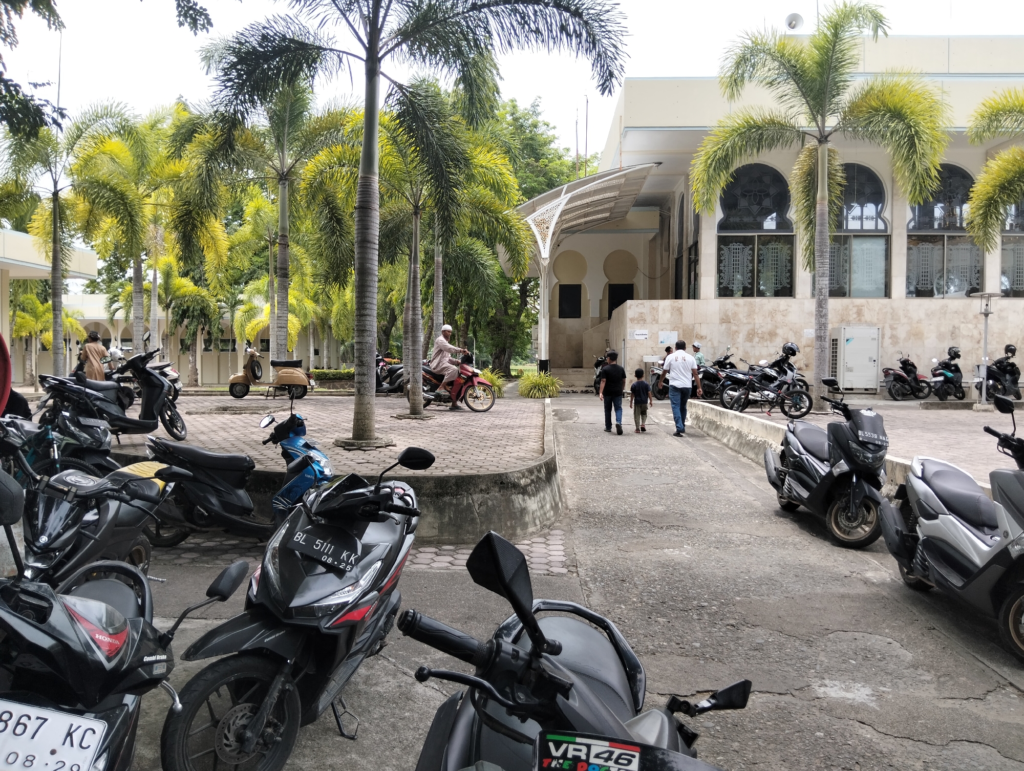 The image depicts an outdoor scene in Tambontunong, Indonesia, characterized by a building with arched windows and a paved area filled with parked motorcycles. Several palm trees with green fronds are scattered throughout the scene, some reaching towards the bright sky.
In the foreground, a cluster of parked motorcycles, primarily scooters, dominates the lower half of the frame. These motorcycles are of various colors including black, grey, and one blue scooter. Some motorcycles have visible license plates with Indonesian characters. A person is seated on a red motorcycle, wearing a white cap and traditional attire.
In the mid-ground, to the right of the building entrance, three individuals are walking away from the viewer. Two appear to be adult males, one in a white shirt and the other in a dark shirt, followed by a child in a green shirt. More motorcycles are parked in this area, including a lighter colored scooter and several black scooters.
The building itself is light-colored with a cream-colored facade and a prominent roof structure extending outwards, supported by pillars. Decorative elements with intricate patterns are visible on the facade and over the entrance. The overall environment suggests a public space, possibly outside a mosque or community center, given the architectural style and presence of parked vehicles. The ground is paved with rough concrete and interlocking bricks in some areas.