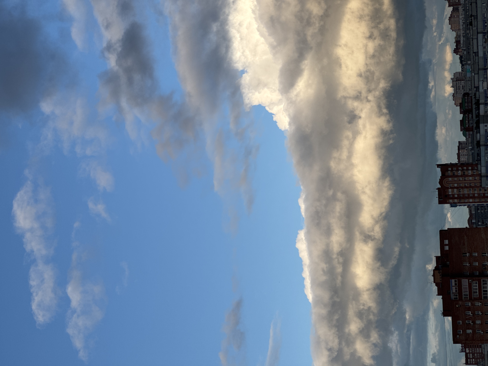 The image shows a view of the sky in an urban setting, with multi-story brick buildings on the right side of the frame. The sky is primarily blue, with scattered small, wispy clouds on the left. A large band of dark, billowing clouds is centrally located, appearing vertical in the frame. These clouds are partially illuminated, exhibiting white and grey tones. The overall impression is one of an approaching storm or significant weather system.