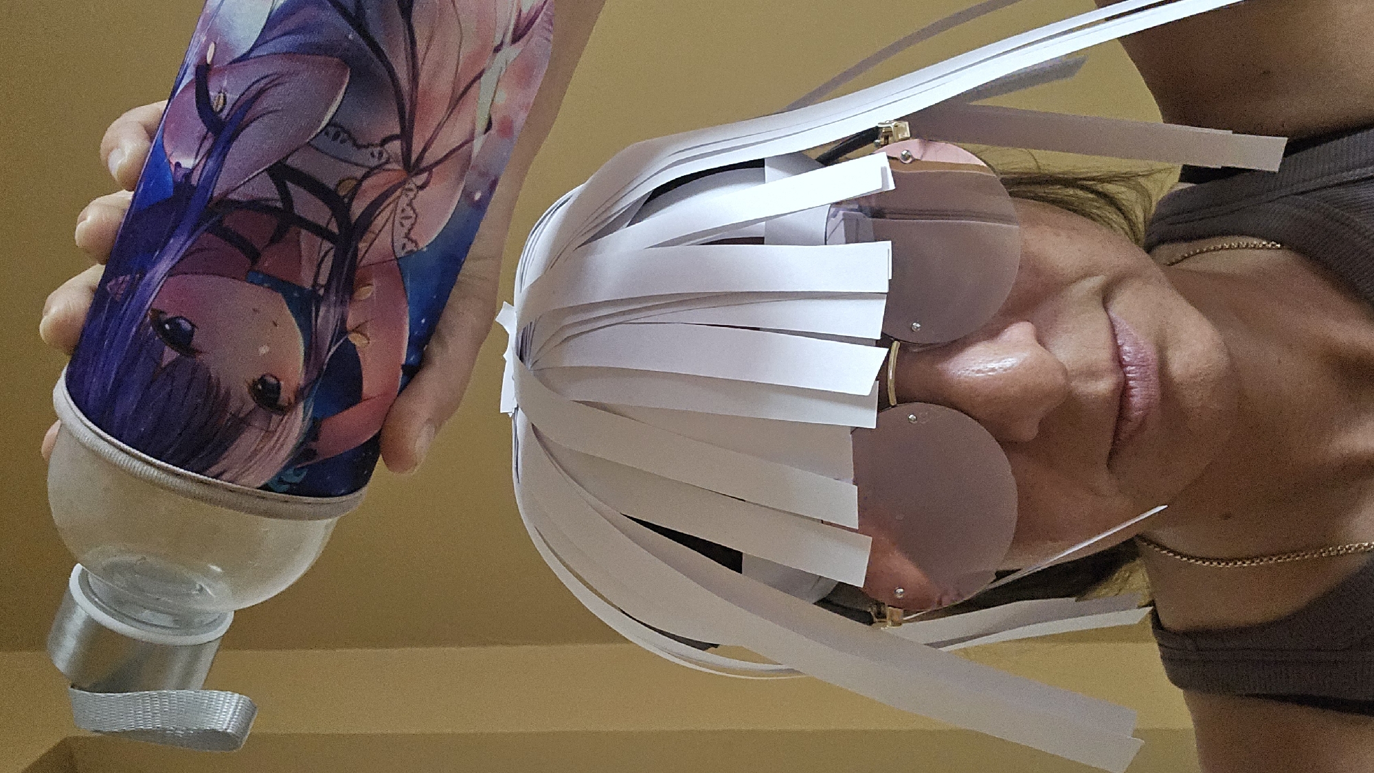 A close-up medium shot captures a person wearing large, pink-tinted, rimless sunglasses and a homemade wig crafted from white paper strips. The wig is attached to the person's head, with the paper strips hanging down like hair. Above the person's head, a hand holds a spray bottle adorned with an anime-style illustration of a young girl. The background is a plain, light brown wall. The scene appears to be indoors.