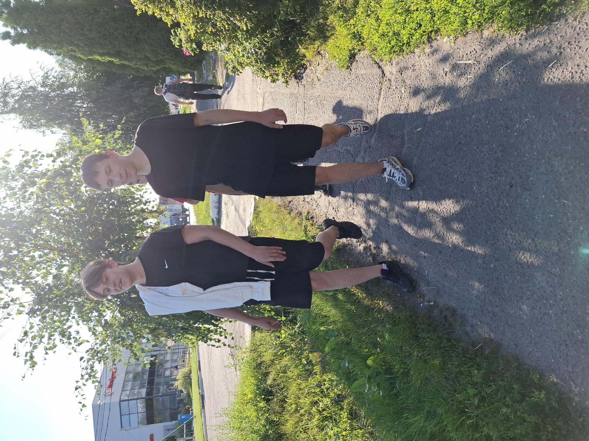 Two young men are walking on a paved pathway in front of a building and trees. The man on the left is wearing a black t-shirt, black shorts, and black sneakers. He has a white jacket draped over his shoulders. The man on the right is wearing a black t-shirt and black shorts and white and gray sneakers. A third person, wearing a black backpack, is walking away from the camera on the pathway in the background. The scene appears to be in an urban or suburban setting, with a street and grassy area to the left of the pathway and lush green bushes and trees to the right. The sun is shining, casting shadows on the pathway. The building in the background has a modern glass facade and a sign with a red "5" logo.
