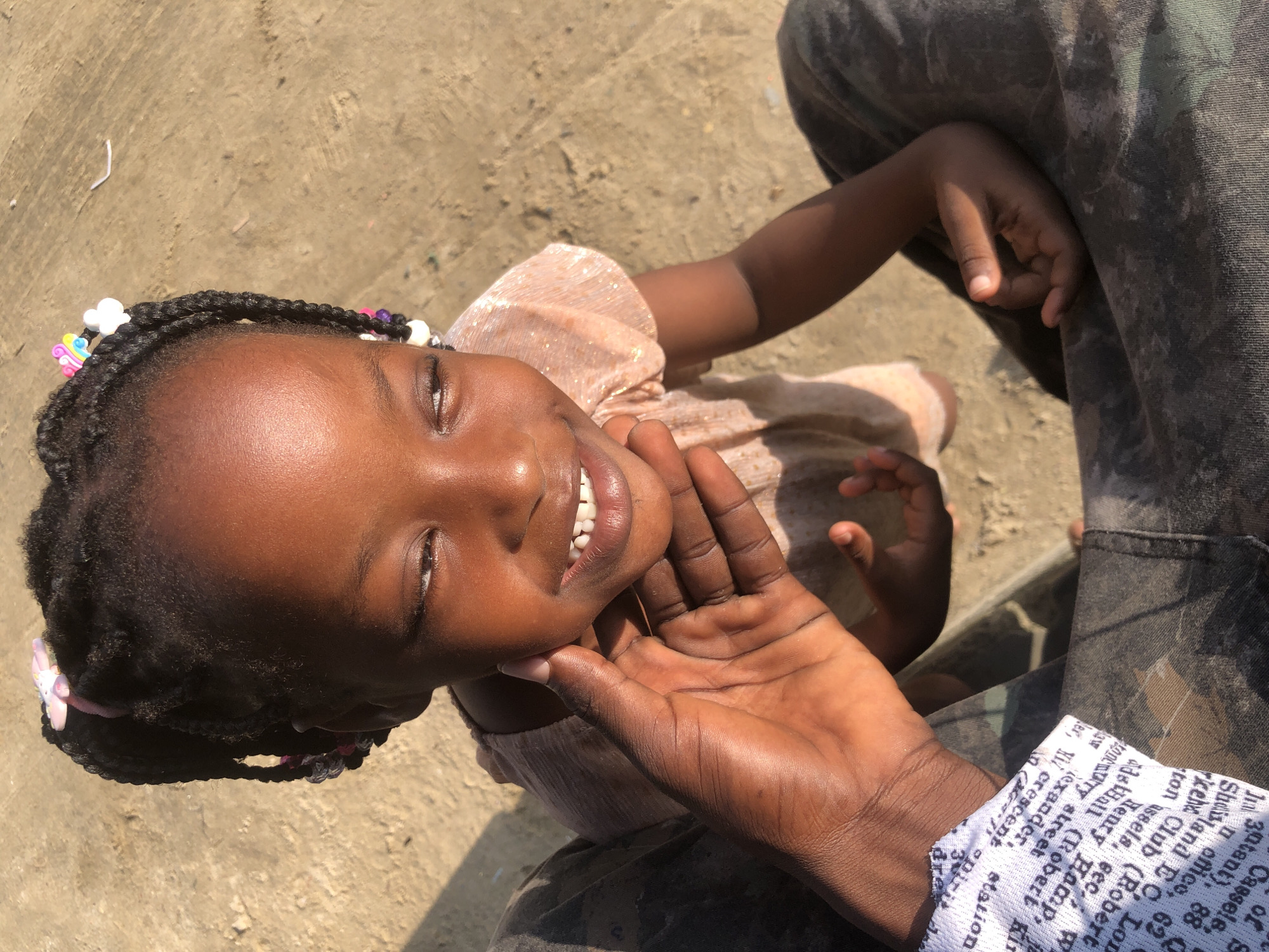 This media file features a close-up, overhead shot of a young Black girl with braided hair adorned with colorful beads and charms, including what appears to be a unicorn charm. She is looking upwards with a bright, joyful expression, her eyes are partially closed, and she is smiling, revealing her teeth. Her chin is being gently held by an adult's hand, whose skin tone suggests they are also Black. The adult is wearing camouflage-patterned pants. The girl is wearing a light, shimmering peach-colored dress. The background is a sandy or dusty ground, indicating an outdoor setting. The lighting suggests it is daytime, likely sunny, casting shadows. The image is framed such that the girl's face and upper body are the primary focus, with the adult's hand and lower body providing context.
Visible text, potentially from the adult's clothing cuff or a piece of paper nearby, includes fragments of what seems to be an address or a list of names and places. Some discernible words and phrases from the OCR are: "Cassels," "office," "street," "crescent," "station," and names like "Robert" and "Henry." This text does not directly relate to the girl's immediate activity but might offer clues about the environment or the adult's belongings. The overall mood is one of tenderness and happiness, captured in a candid moment outdoors in what is identified as Ado, Nigeria.