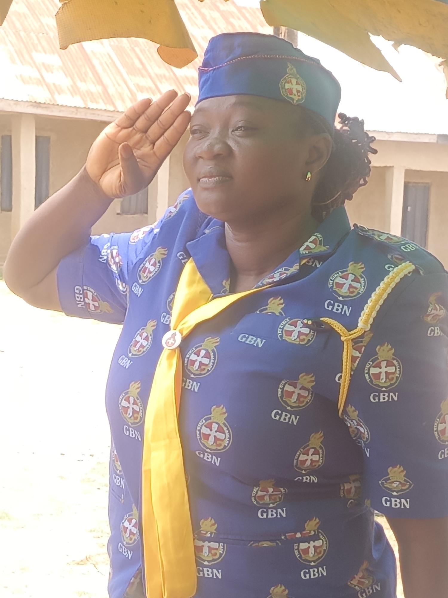A middle-aged African woman in a blue uniform and cap salutes outdoors. The uniform is patterned with the emblem of the Girls' Brigade Nigeria (GBN). She wears a yellow neckerchief secured with a circular clasp. A yellow and white cord is visible on her right shoulder. The background includes a building with a corrugated metal roof and a white exterior wall. The photograph appears to be taken in Akwanga, Nigeria.