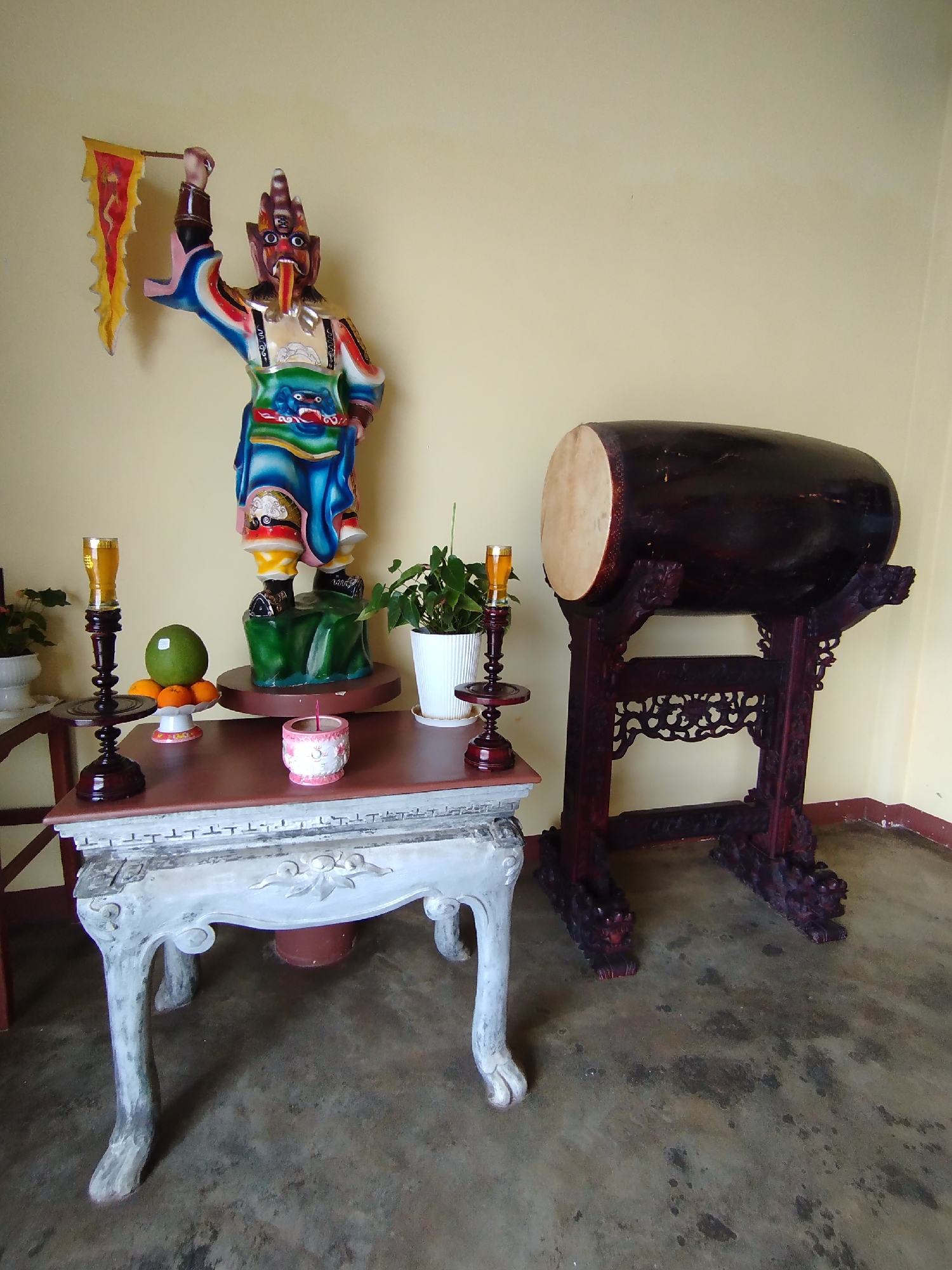 Captured in the city of Phú Lộc, Vietnam, the image presents a scene of religious or cultural significance. A colorful statue, possibly representing a deity or mythical figure, stands atop a small, ornate table. The statue holds a flag in its raised right hand.
On the same table, offerings such as fruit and lit candles are arranged, along with a small potted plant. Another, taller plant stands in a white pot on the table's corner. To the right of the table, a large, dark brown drum rests on an intricately carved stand.
The background is a simple, light yellow wall, and the floor is a mix of grey and brown hues with mottled patterns. The image suggests an indoor setting, possibly a temple or place of worship. The lighting is soft and even, indicating natural light or well-distributed artificial light.