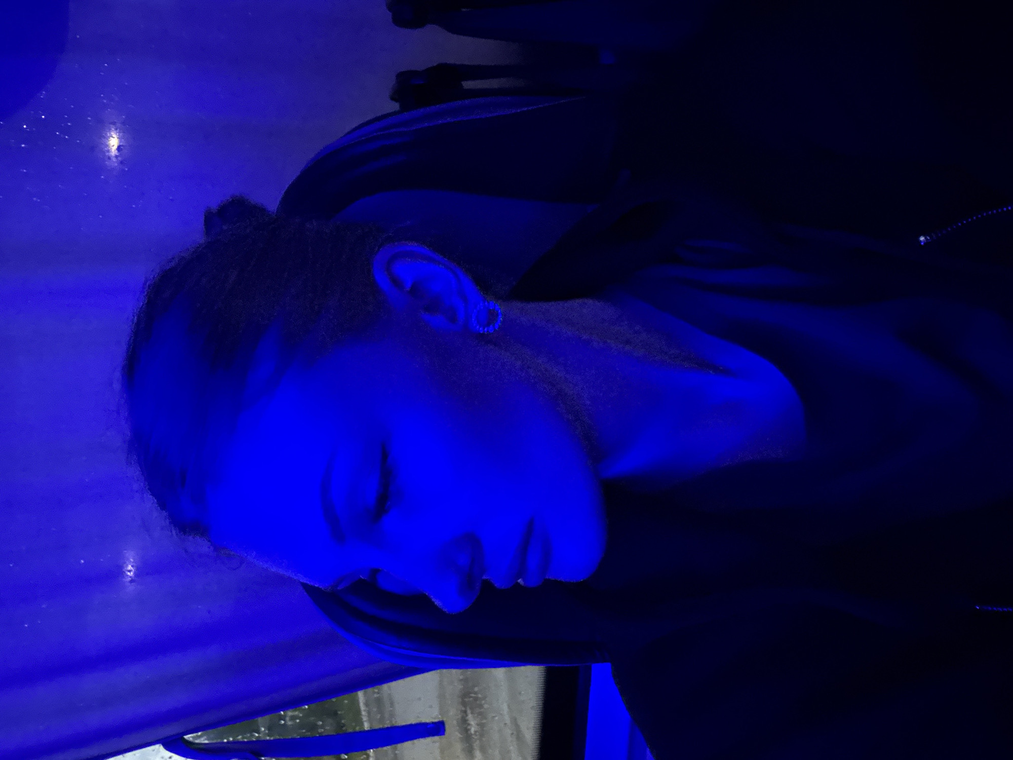 A medium shot photograph captures a young woman in profile, illuminated by a strong blue light. She is seated, likely in a vehicle, as indicated by a window with raindrops visible to her left. Her head is turned to the left, and her gaze is directed downward and away from the camera. She has dark hair pulled back and wears a subtle earring. The blue lighting casts deep shadows and highlights her facial features, including her nose, lips, and cheekbone. The background is predominantly dark, with hints of the interior of a vehicle, possibly seats and a curtain or blind. The overall ambiance is moody and introspective due to the lighting and the woman's posture. The scene is set in Kazan, Russia.
