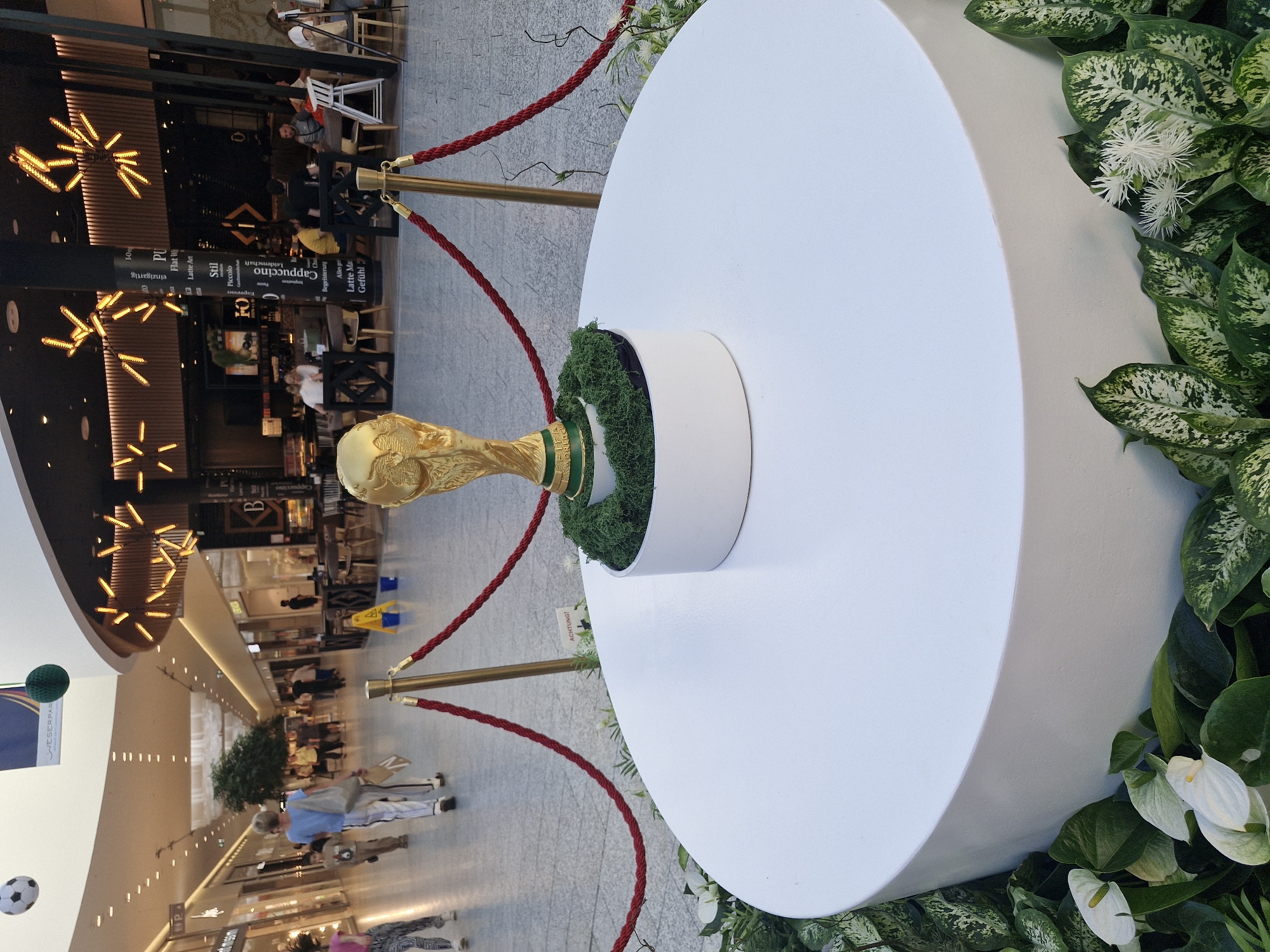 The image depicts a FIFA World Cup trophy on display in a bright, modern indoor shopping mall. The trophy is gold, sits on a white cylindrical pedestal, and is surrounded by artificial moss and green plants. Red ropes with gold stanchions cordon off the display area. In the background, people are walking through the mall, some are seated at tables in a cafe, and decorative lighting fixtures hang from the ceiling. The mall appears to be clean and well-maintained, with polished floors and storefronts visible in the distance. A portion of a sign is visible that reads "LÜNESER PARK" which indicates the location is likely within or associated with Lüneburger Heide region of Germany. However, it does not specifically identify the city as Bremen.