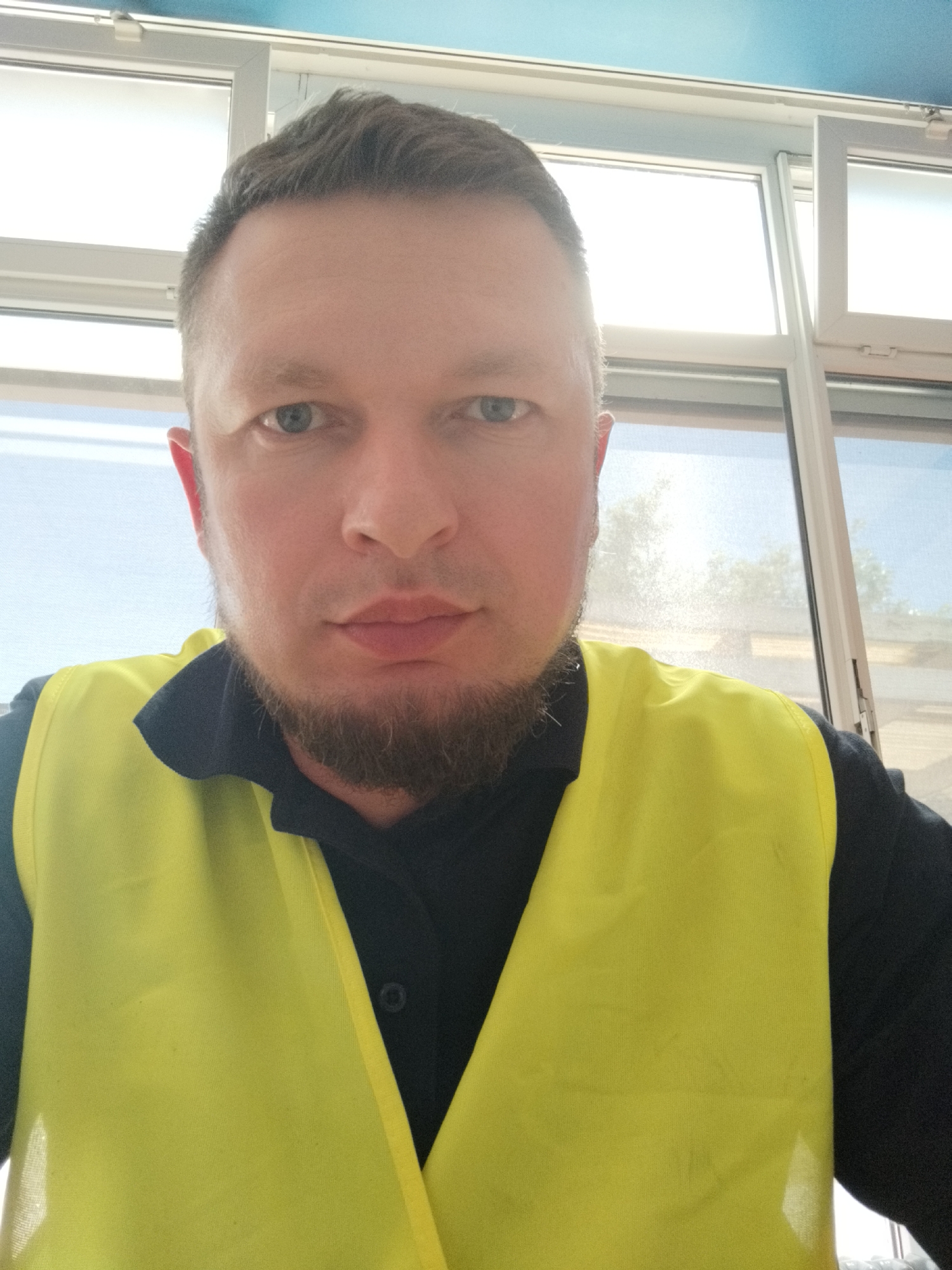 A man with short brown hair and a short beard is shown in a close-up selfie. He is wearing a dark shirt and a bright yellow safety vest. He is looking directly at the camera with a neutral expression. The background appears to be an office or indoor setting with large windows showing a bright outdoor environment with trees. The exact location in Bremerhaven, Germany, is not determinable from the image.