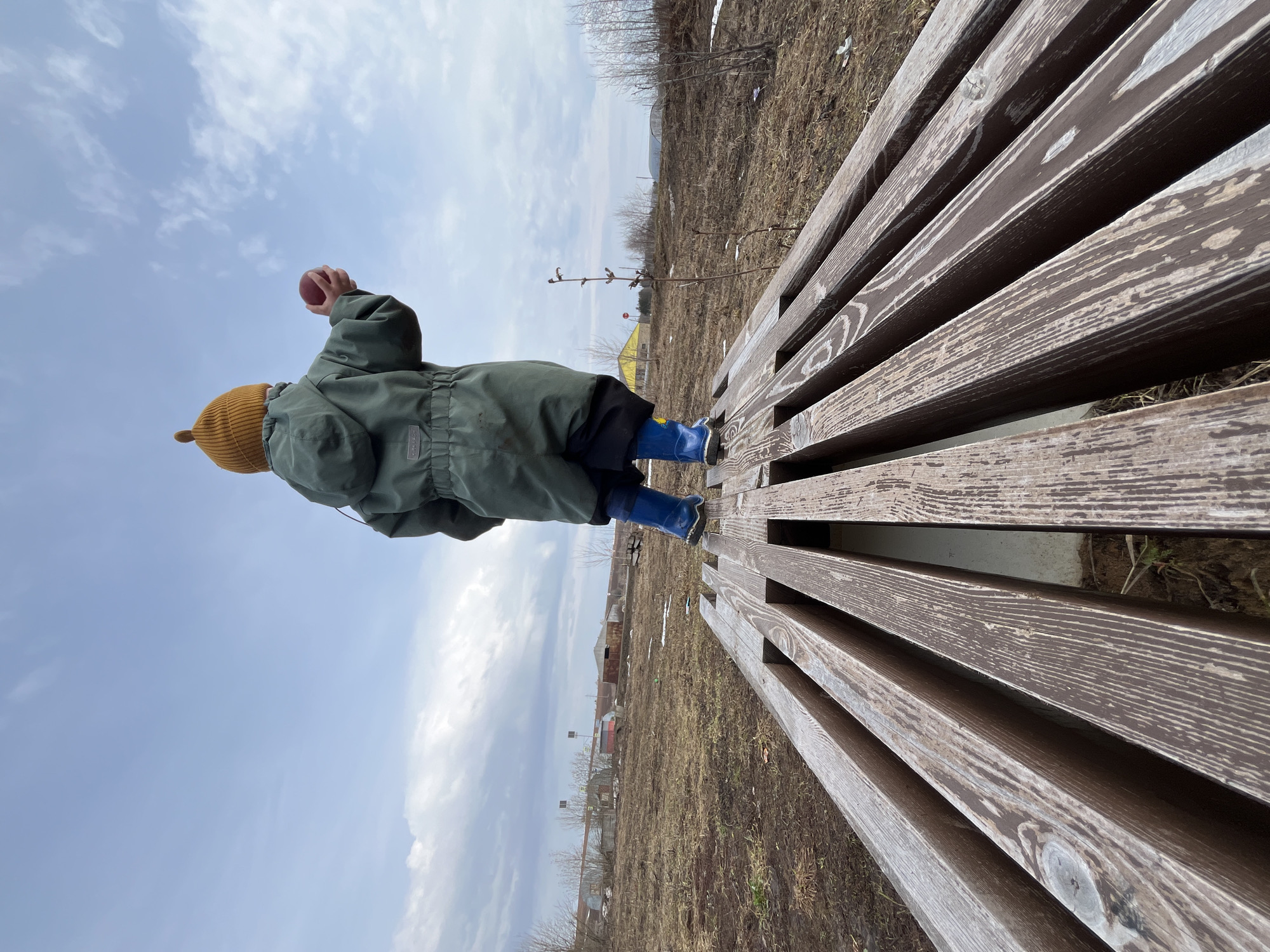 A child wearing a mustard-colored hat, green jacket, and blue boots walks on a wooden bench in Павловка, Russia. The ground is mostly bare, with some patches of snow. In the background, there are trees, houses, and a cloudy sky. The child is holding a small, round object in their hand.