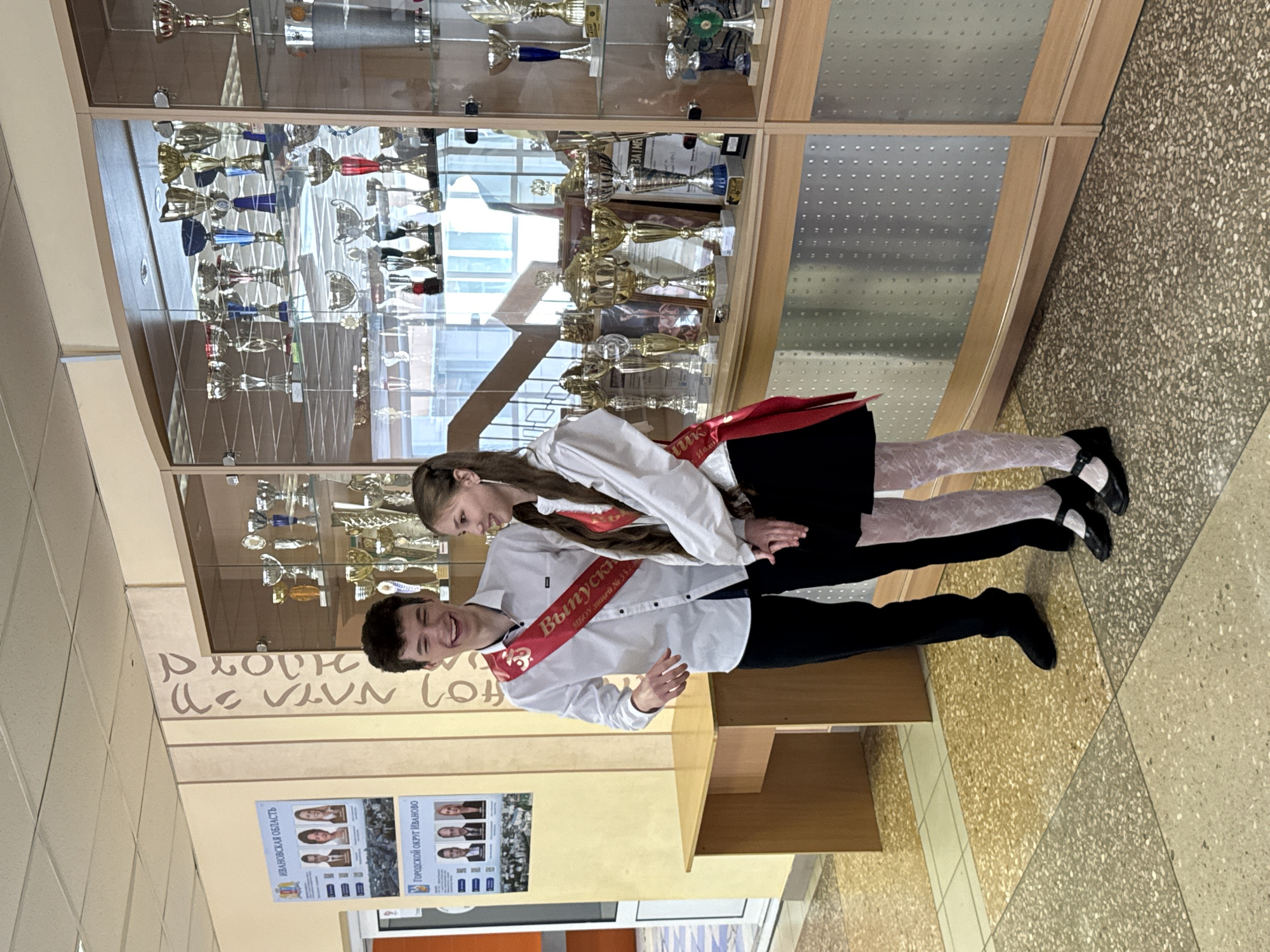 Two young people, a male and a female, stand side-by-side in a hallway. Both are wearing white shirts and graduation sashes with Russian writing. The male is on the left with black trousers and shoes. The female wears a black skirt, patterned white tights, and black shoes. They are positioned in front of a display case full of trophies. Behind them is a wall with the words of the city of Ivanovo written on it. A poster is also on the wall below the writing that says "Ivanovo Region" and has photos of people on it. The floor is tile and the lighting is bright and fluorescent.