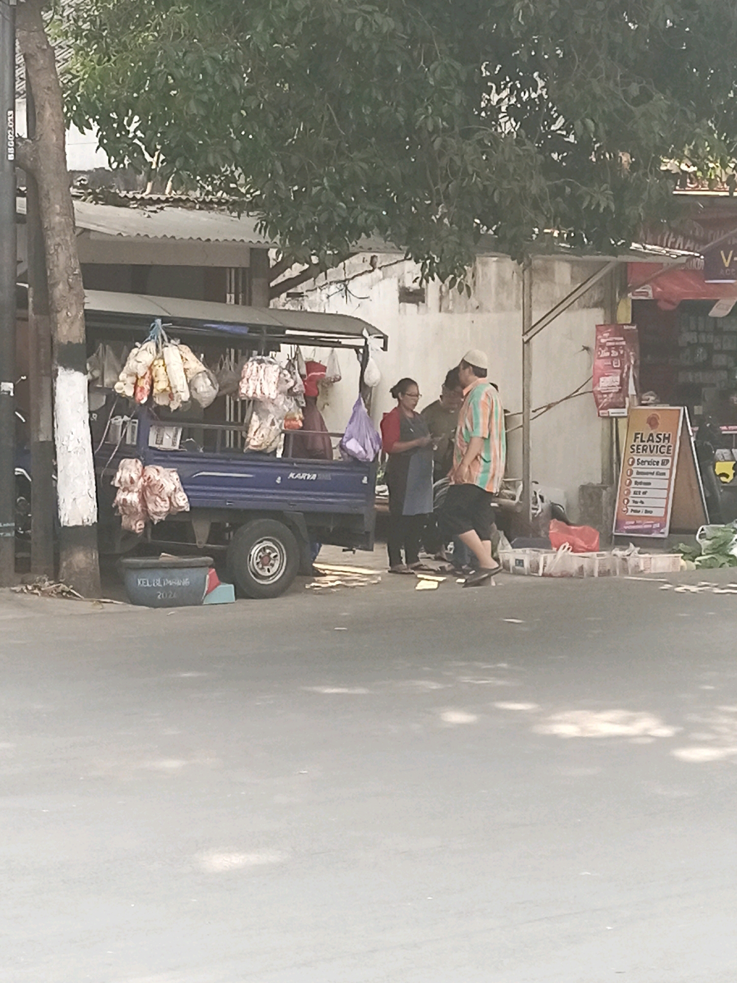 The image depicts a street scene in Malang, Indonesia, likely on a sunny day. In the foreground, the asphalt street is visible, with dappled sunlight creating patterns. A blue truck, possibly a pickup truck, is parked on the side of the road. Bags of goods are hanging from the side of the truck, and a blue plastic tub with white lettering is placed in front of it.
Behind the truck, a group of people are gathered. One woman, wearing a red shirt and a blue apron, appears to be interacting with a man wearing a striped shirt and a white cap. Another man is partially visible behind them. The background shows a building with a corrugated metal roof and a white wall. A sign with the text "FLASH SERVICE" and a list of services is propped up against the building. Various other items, including plastic containers and what appear to be fresh produce, are scattered on the ground near the people and the sign. A mature tree with dense foliage shades part of the scene. The overall atmosphere suggests a marketplace or roadside stall.