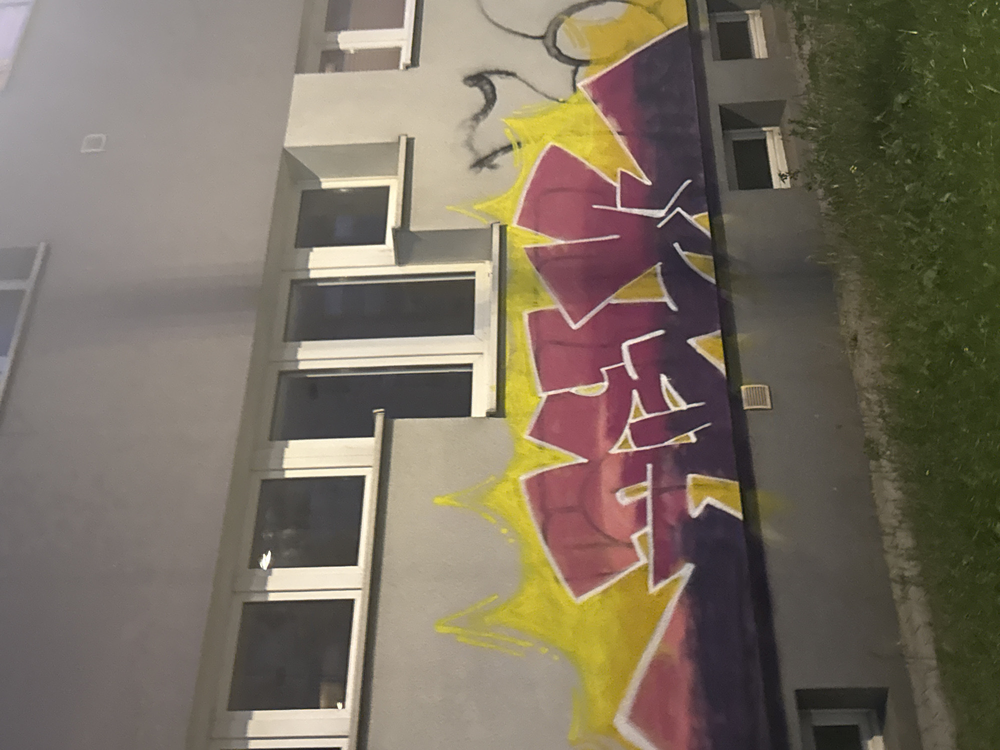 The image shows a section of a building's exterior wall adorned with graffiti. The building has a gray facade and several windows with white frames. The graffiti features large, stylized letters in purple and pink with white outlines, set against a bright yellow background that appears to be spray-painted in a jagged, flame-like pattern. Below the graffiti, the wall transitions to a darker gray, and there are small windows or vents visible near the ground level. The bottom of the image shows a strip of green grass. The overall scene appears to be an outdoor urban environment, likely at dusk or night, given the lighting.