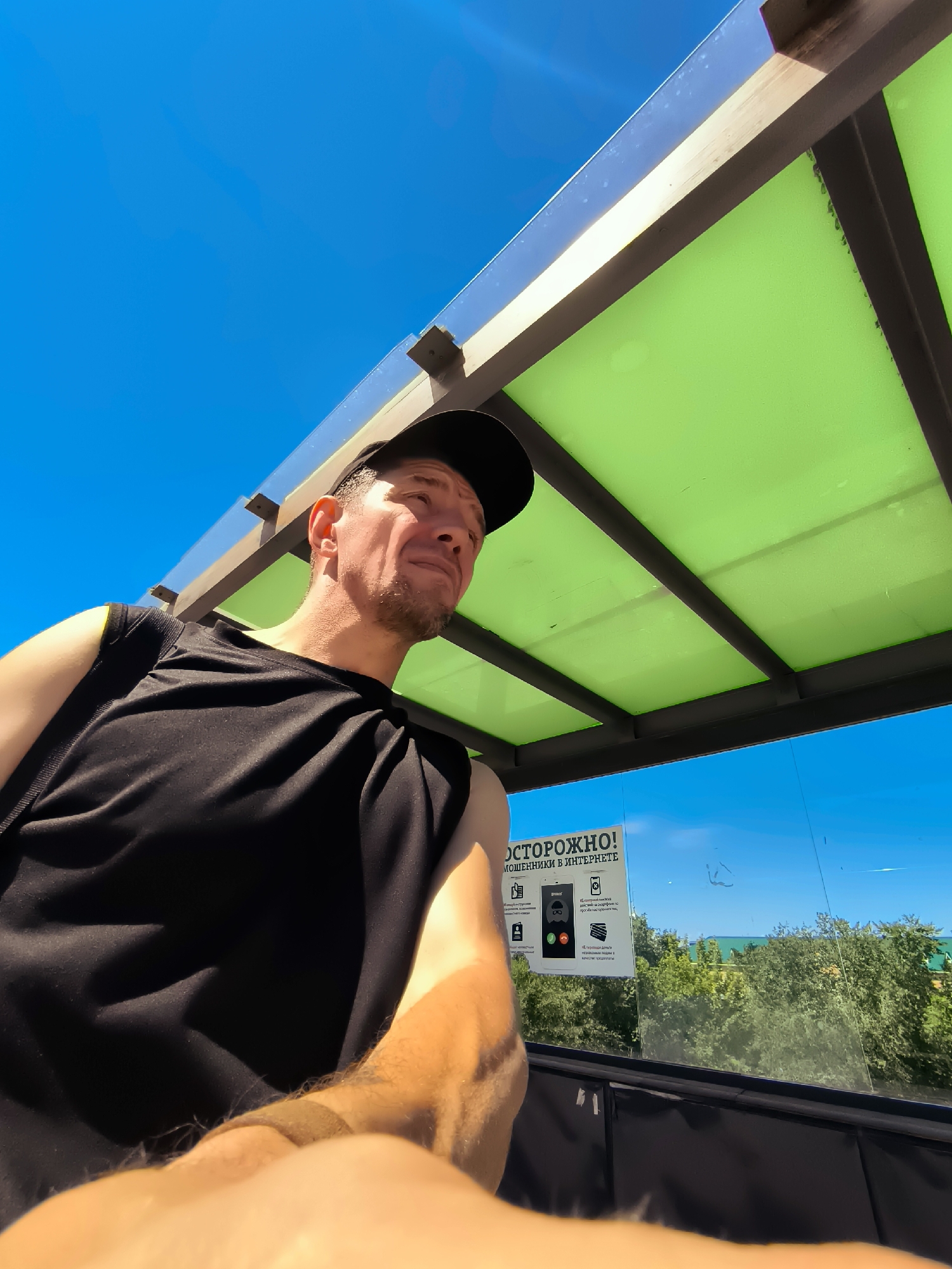 A man wearing a black sleeveless shirt and a black baseball cap is looking up and to the right. He has short brown hair and stubble. He is in what appears to be a covered outdoor seating area, possibly a bus stop or pavilion, with a green translucent roof and a clear glass wall behind him. Outside the glass wall, there is a view of green trees and the blue sea under a bright blue sky. A sign is visible on the glass wall with Russian text and icons. The man's left arm and hand are visible in the foreground. The image is taken from a low angle.
